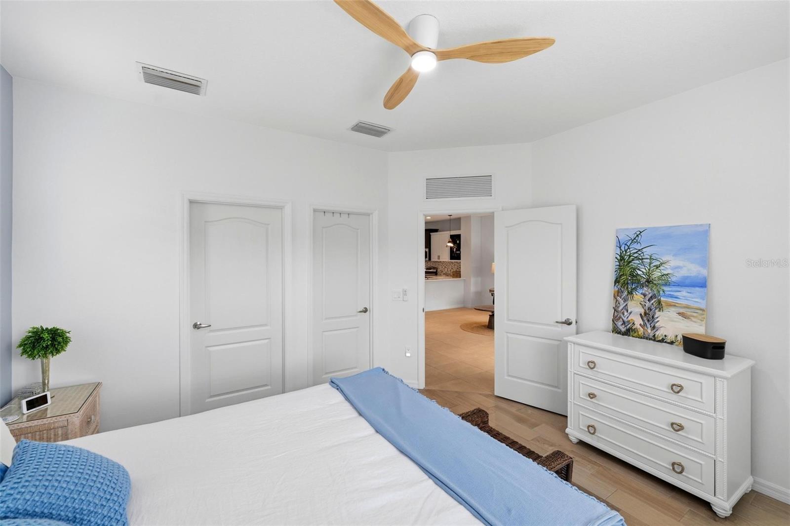 Bright bedroom with a white bed, ceiling fan, and dresser beside an open doorway.