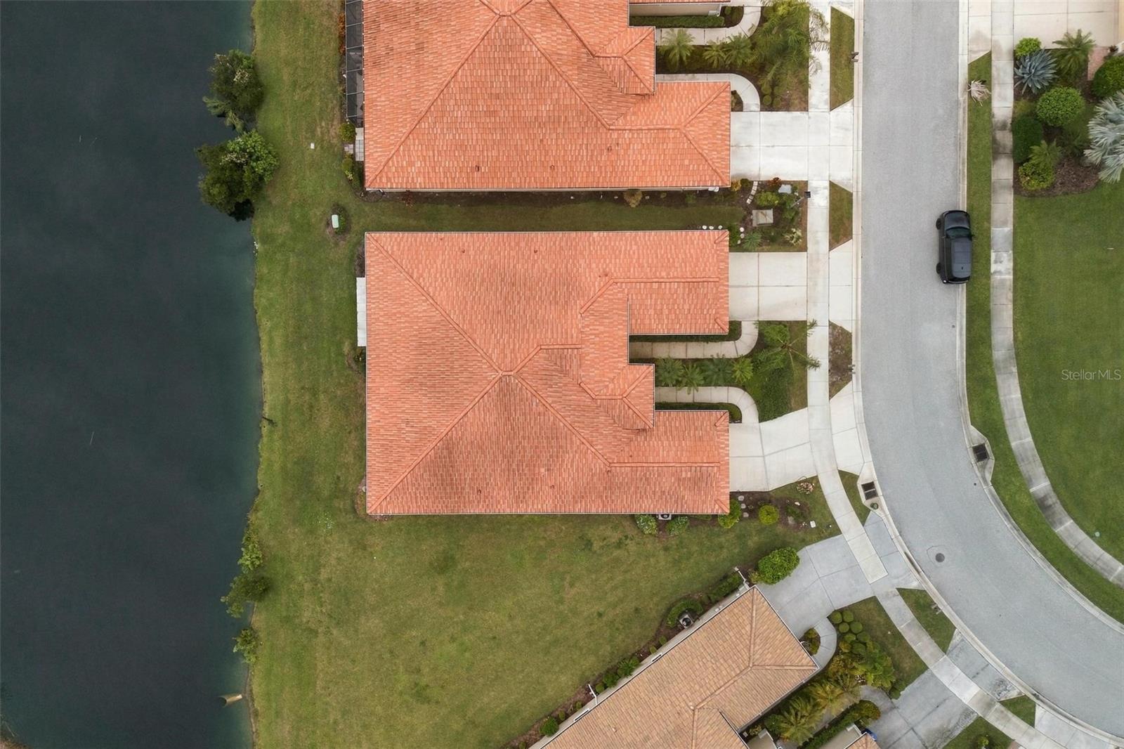 Aerial view of a tiled-roof home beside a pond, with its lawn and driveway visible.