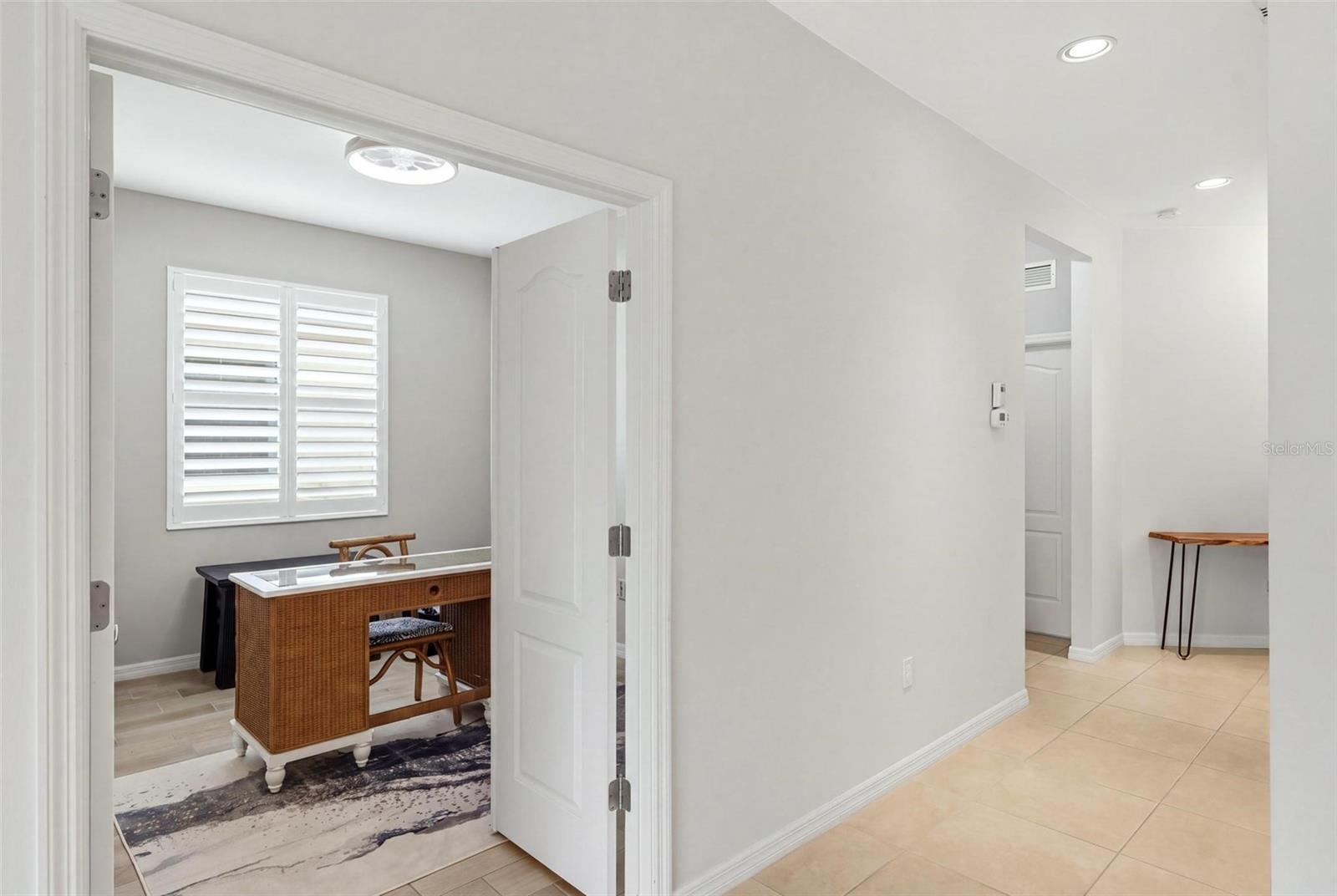 Home office with a wicker desk, plantation shutters, and an open door off a tiled hallway.