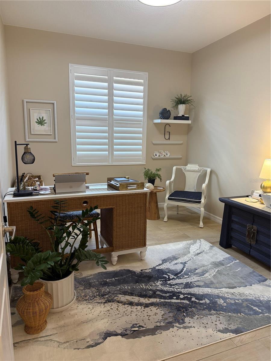 Home office with a wicker desk, white shutters, and a blue-gray patterned rug.
