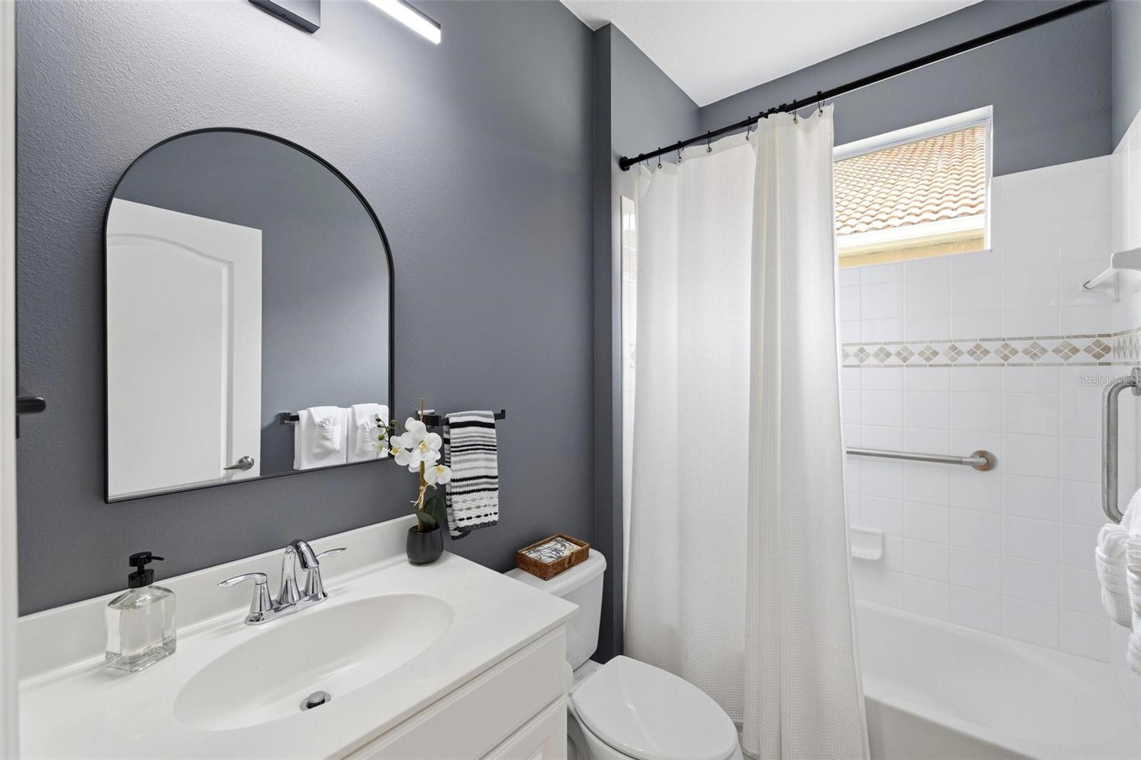 Bathroom with a white vanity, arched mirror, and tiled tub-shower beneath a window.