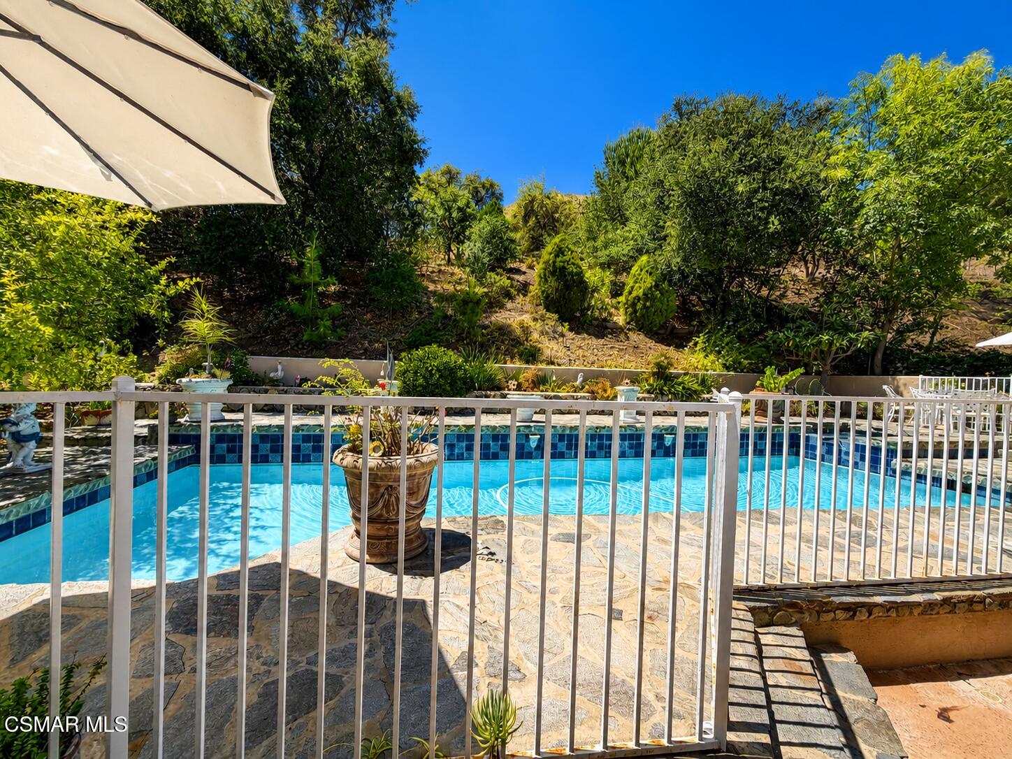 Backyard swimming pool behind a white safety fence, with a stone patio and landscaped hillside.