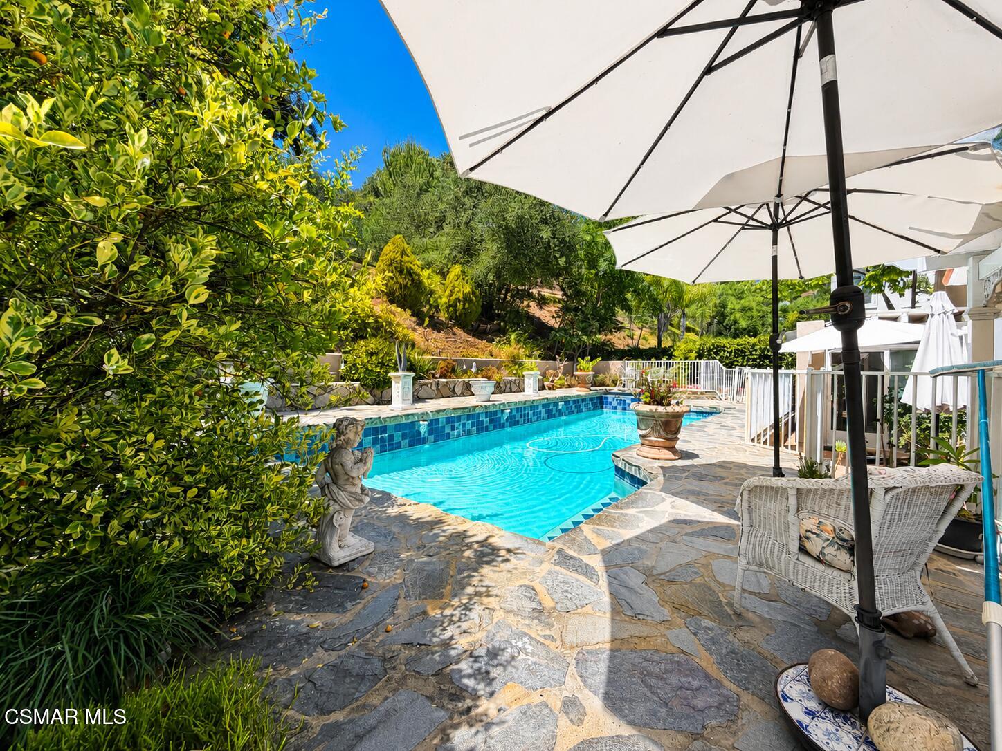 Backyard swimming pool bordered by a stone patio, white umbrellas, and lush greenery.