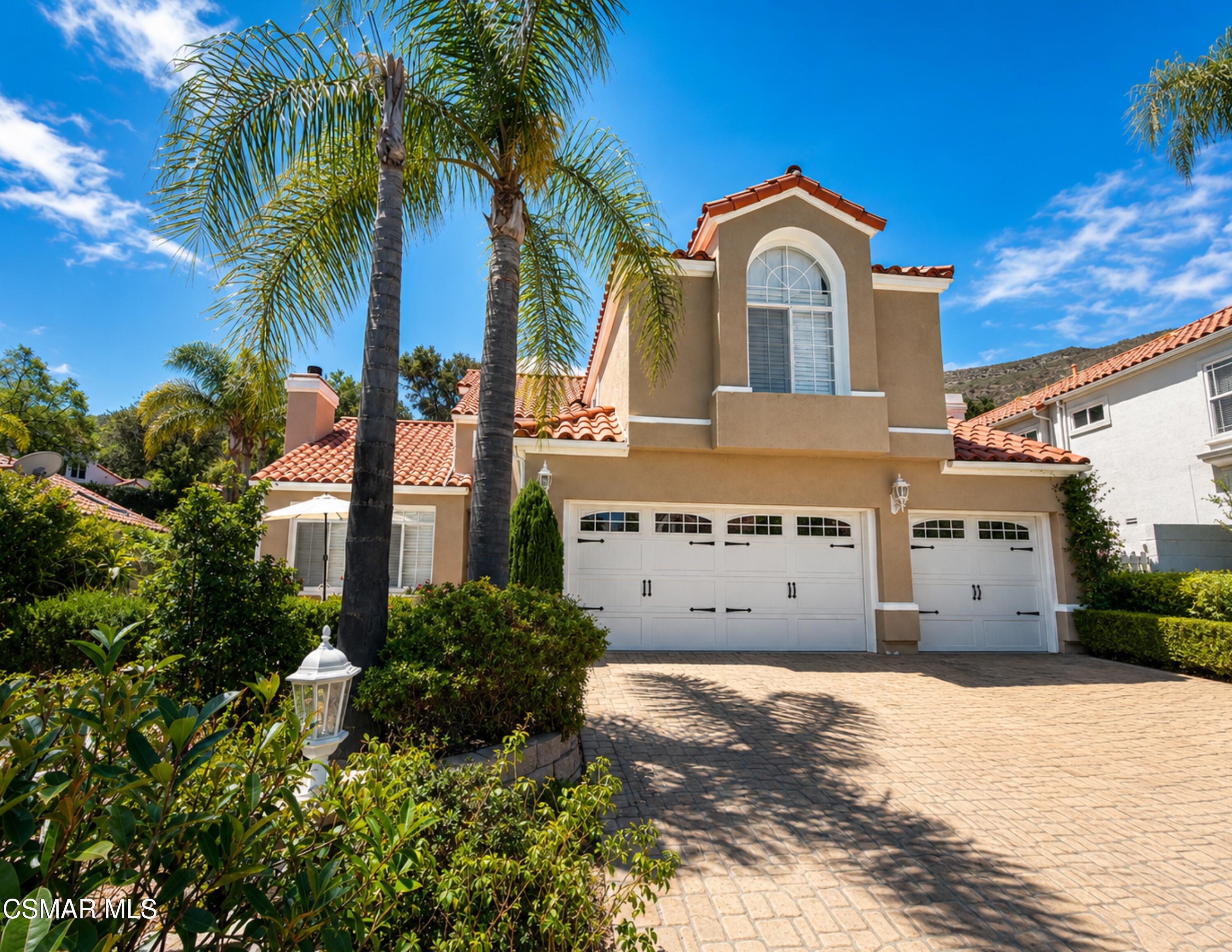 Two-story tan stucco home with red tile roof, arched window, palm trees, and three white garage doors.