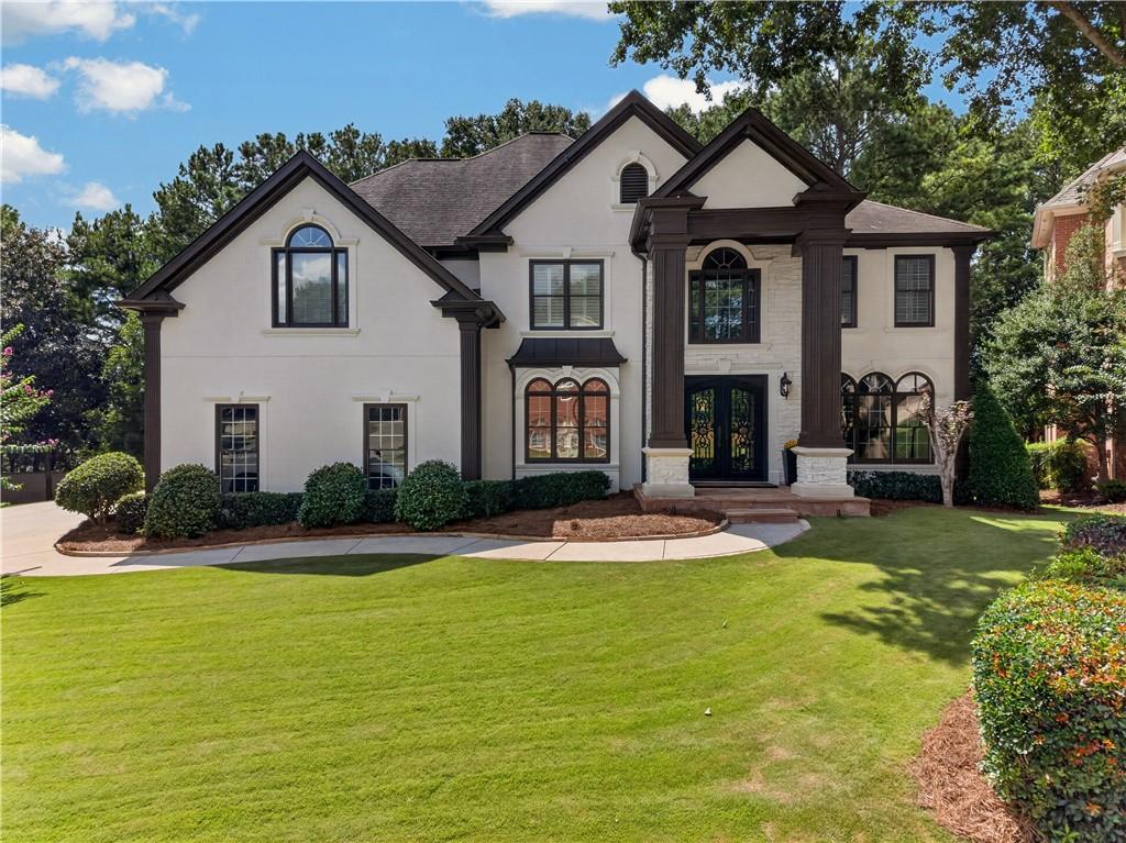 Two-story cream stucco home with dark trim, prominent columns, and a landscaped front lawn.