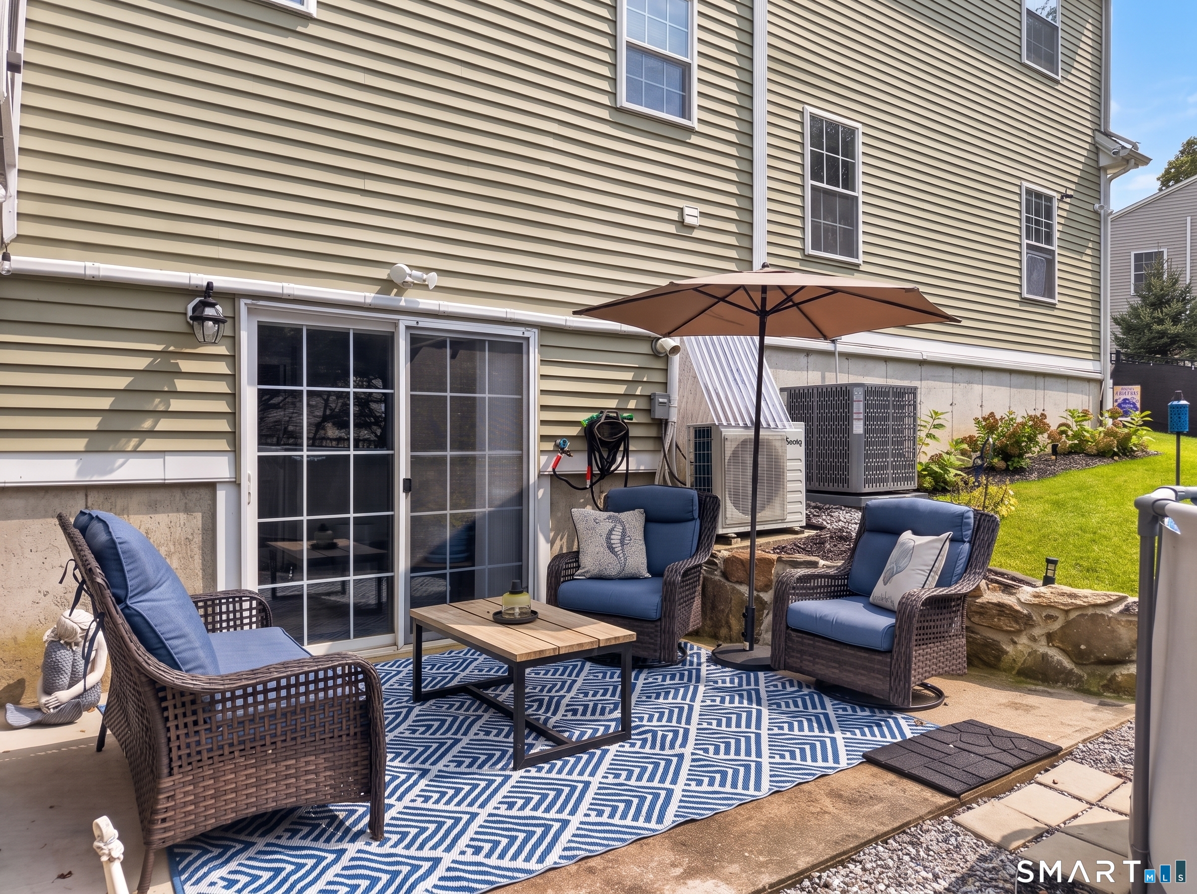 Furnished backyard patio with blue-cushioned wicker chairs, patterned rug, and umbrella beside the house.