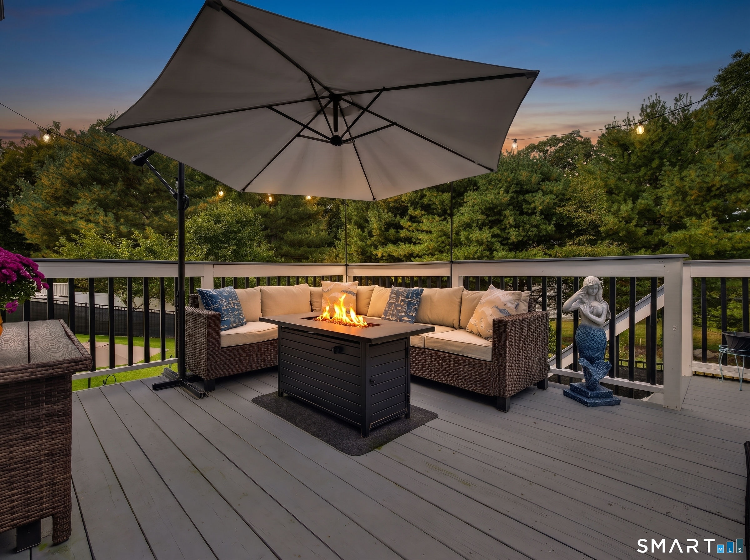 Deck seating area with a large umbrella, fire table, and string lights at dusk.