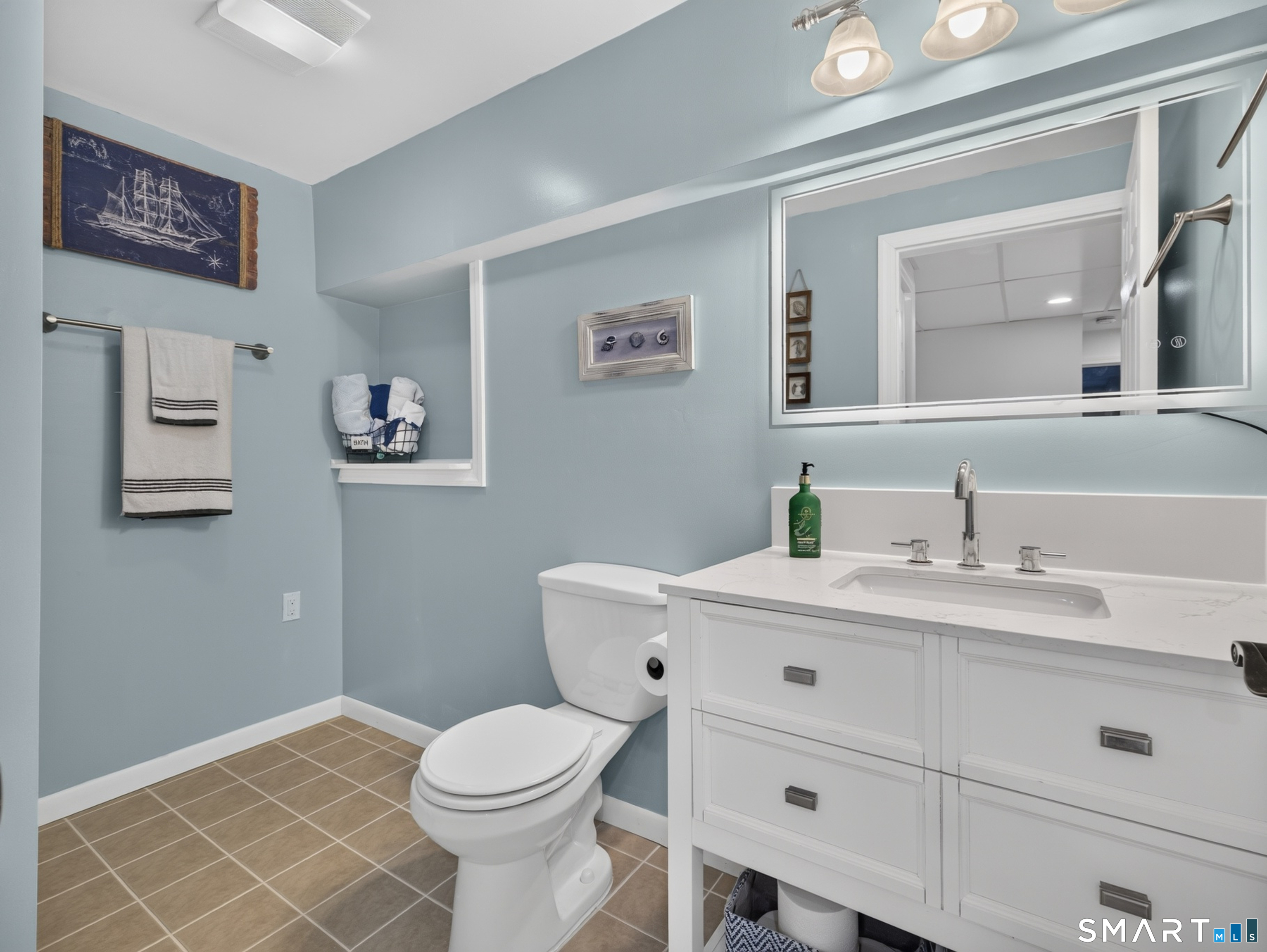 Bathroom with a white vanity and large mirror, toilet, and tan tile floor.