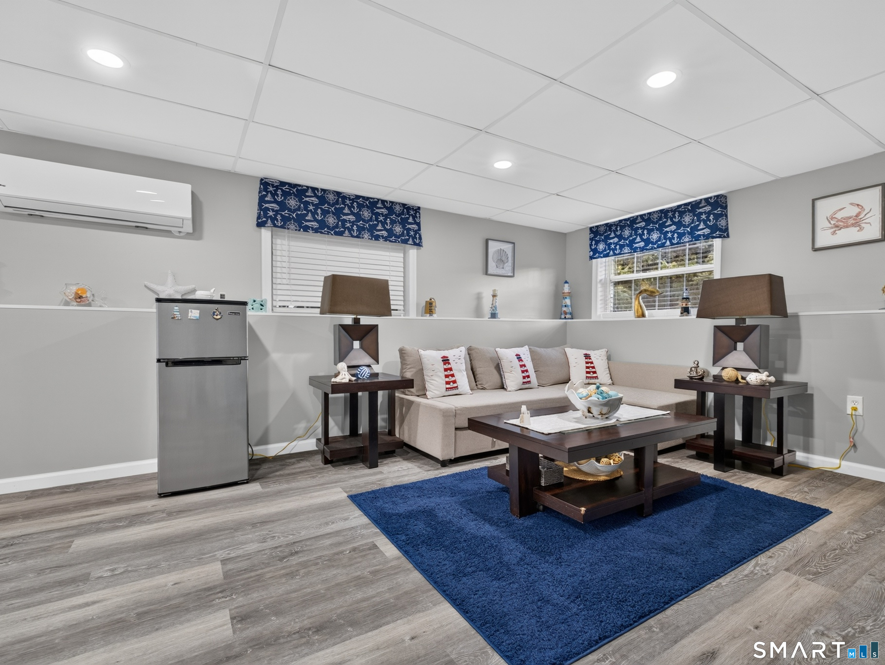 Furnished living room with a beige sectional, nautical accents, blue rug, and gray wood-look flooring.