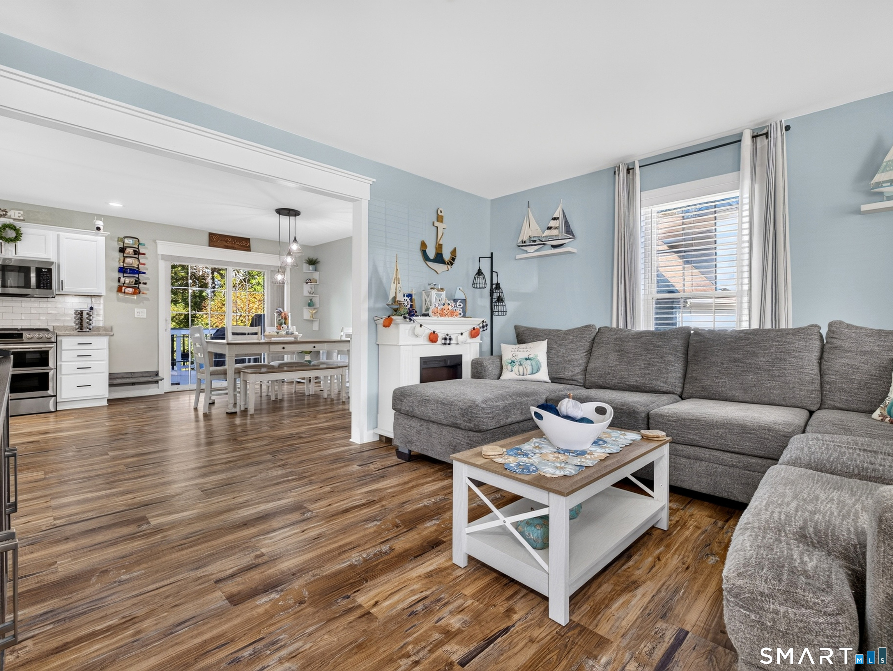 Open living room with gray sectional, fireplace, and adjoining white kitchen and dining area.