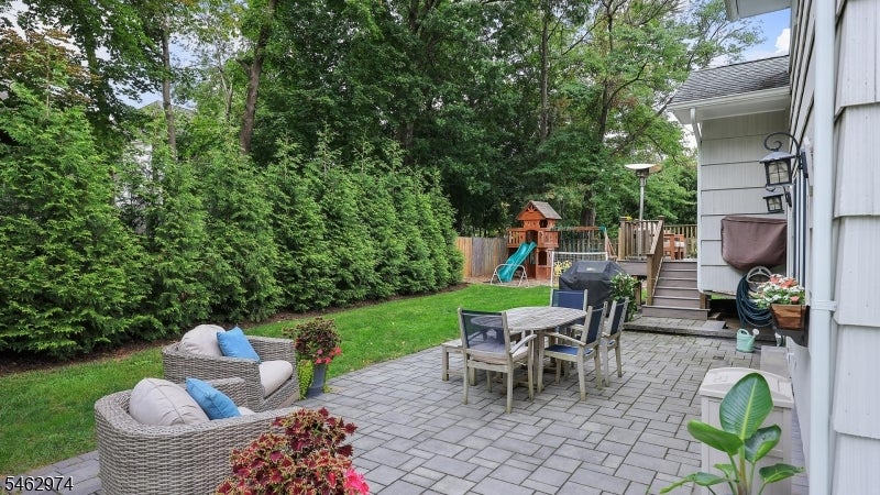 Backyard patio with wicker seating, outdoor dining table, lawn, and a children's playset.
