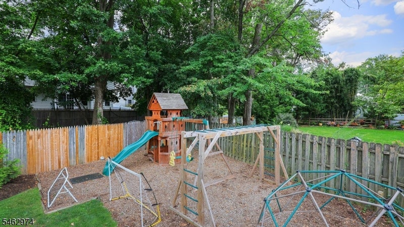 Fenced backyard play area with a wooden playset, teal slide and swings beneath mature trees.