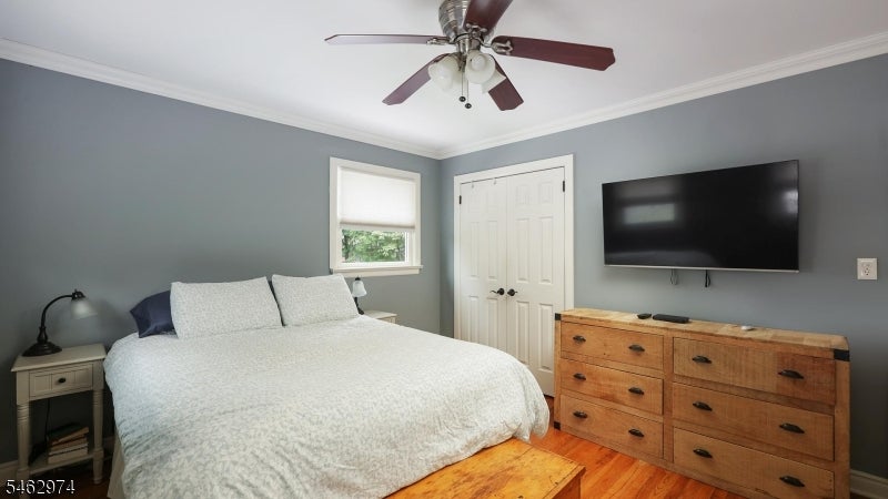 Bedroom with a white bed, wooden dresser, wall-mounted television, and ceiling fan.