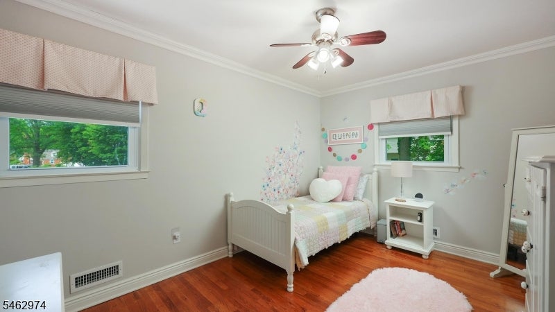 Bedroom with a white bed, pastel pink bedding, hardwood floors, and windows overlooking trees.