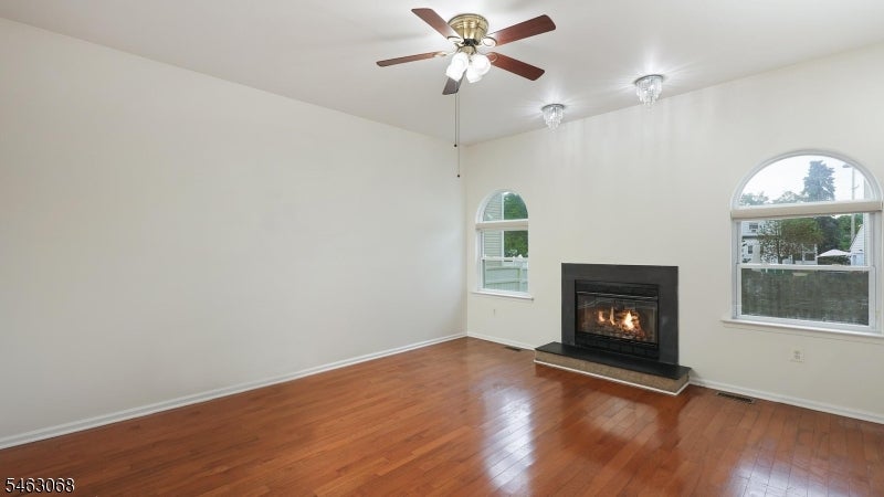 Empty living room with hardwood floors, a lit fireplace, arched windows, and a ceiling fan.