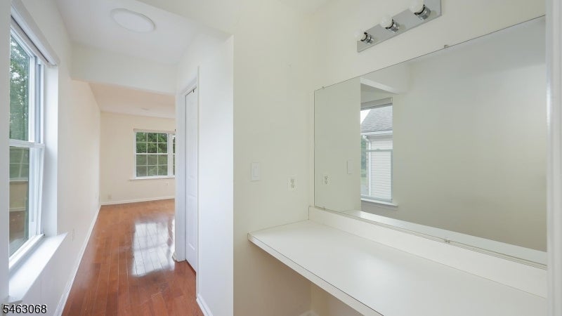 White vanity counter and mirror beside an empty room with reddish hardwood floors and windows.
