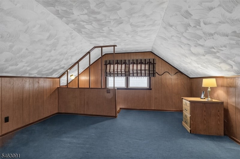 Attic room with sloped ceiling, wood-paneled walls, blue carpet, and a small window.