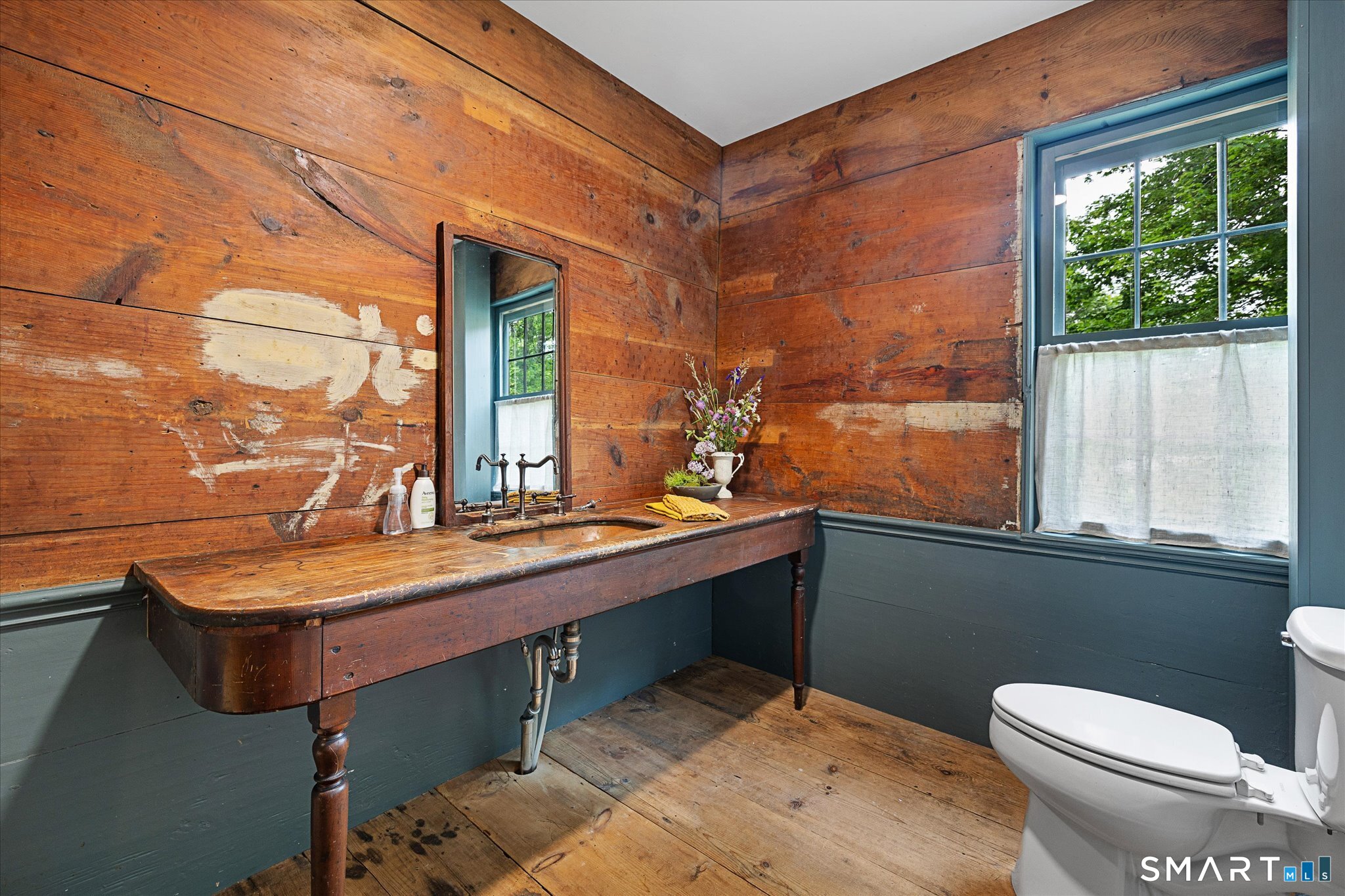 Rustic bathroom with a long wooden sink vanity, weathered plank walls, and a curtained window.