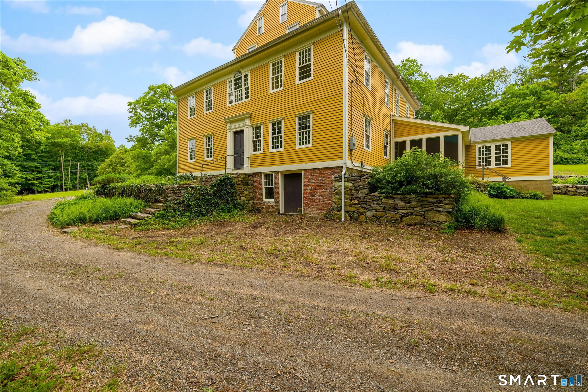 Yellow clapboard house with attached wings, a stone foundation, and a gravel driveway.