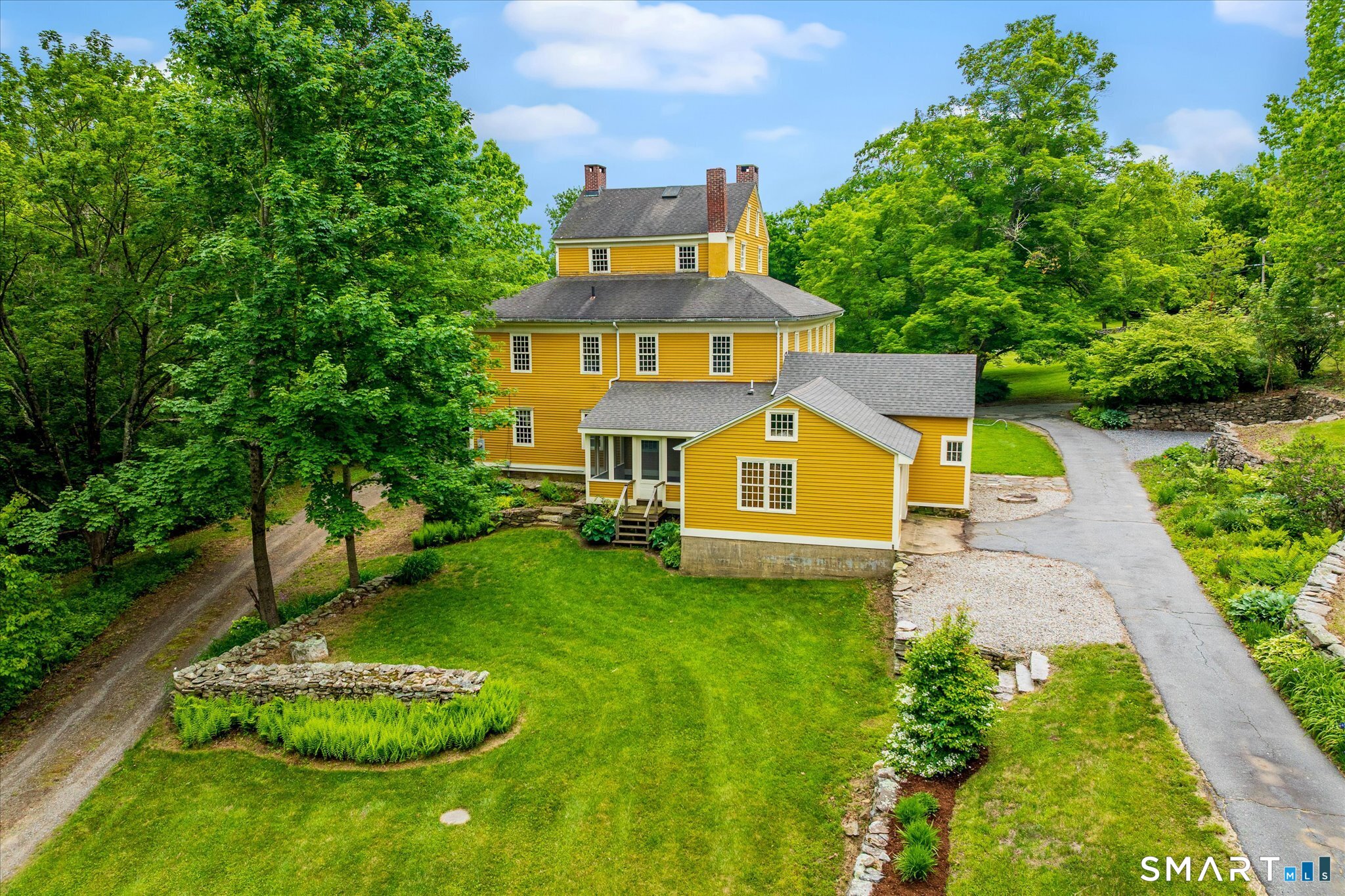 Yellow multi-story house with brick chimneys, landscaped lawn, stone walls, and a curving driveway.