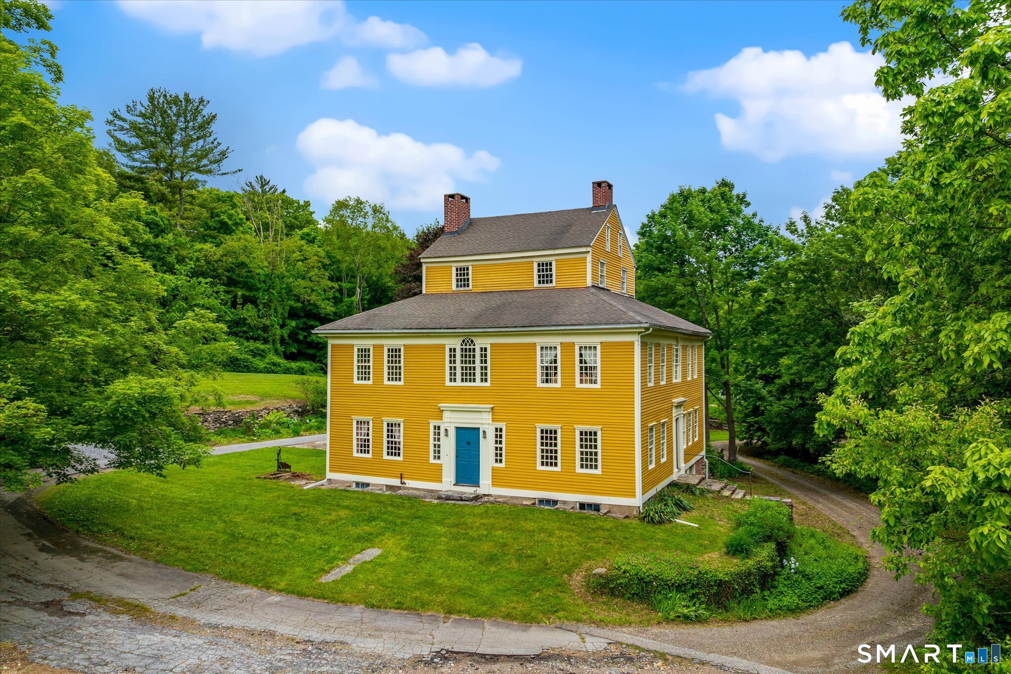 Yellow two-story house with white trim, a blue front door, and brick chimneys amid leafy trees.