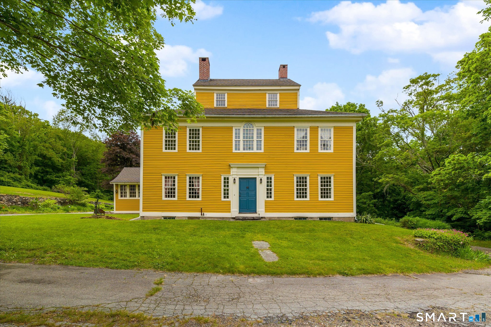 Yellow two-story house with white trim, blue front door, and brick chimneys, surrounded by mature trees.