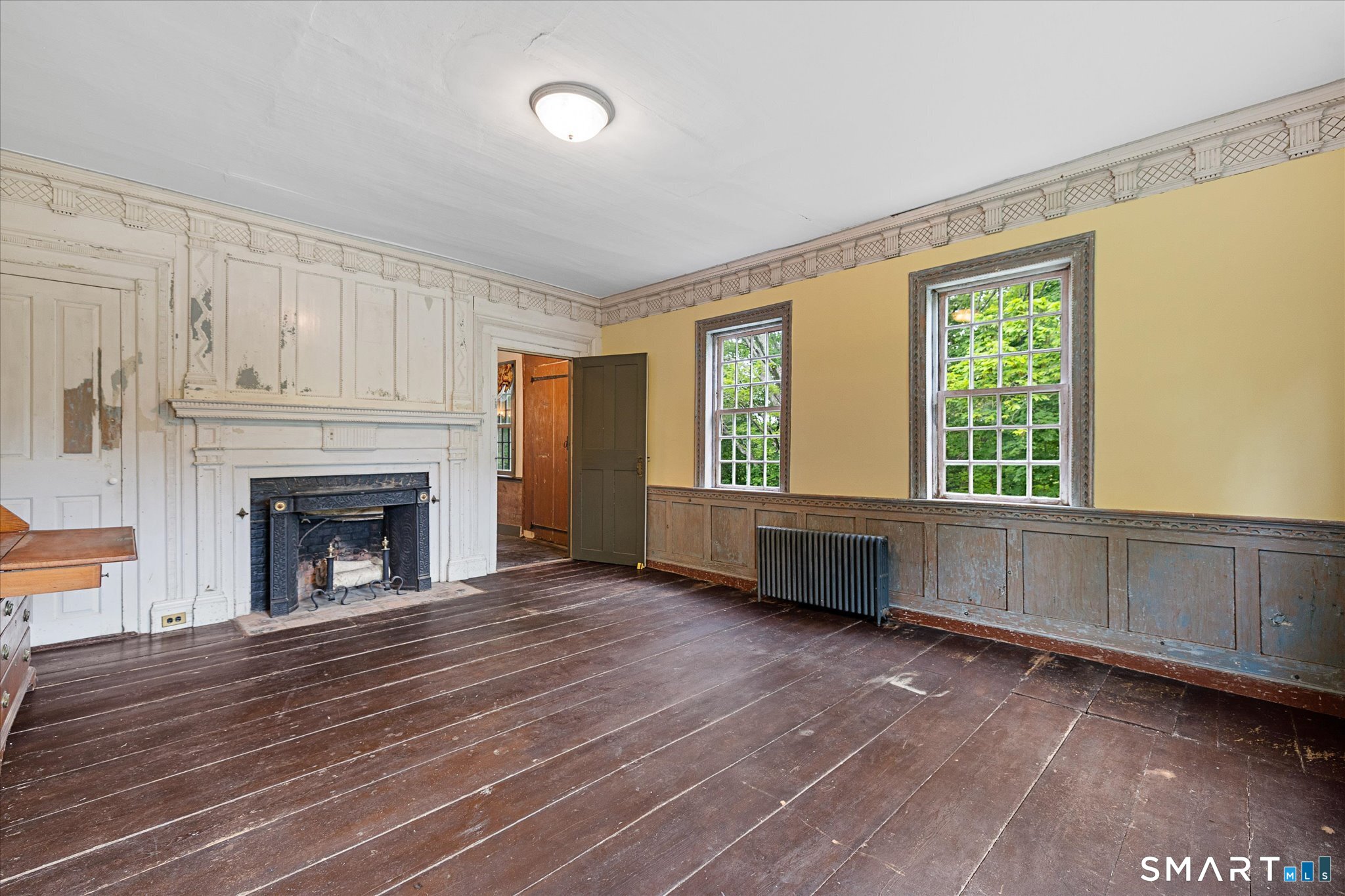 Empty room with an ornate fireplace, historic wood paneling, wide-plank floors, and windows overlooking greenery.