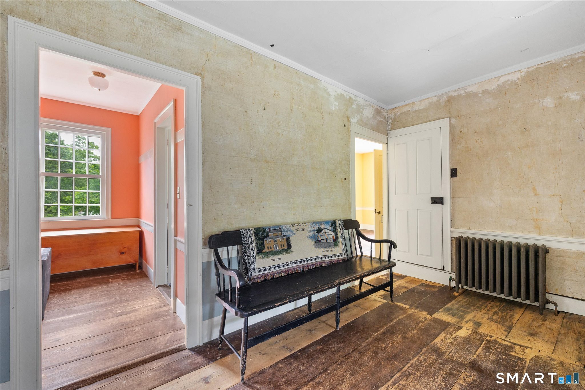 Historic hallway with worn plaster walls, wide plank floors, a black bench, and a cast-iron radiator.