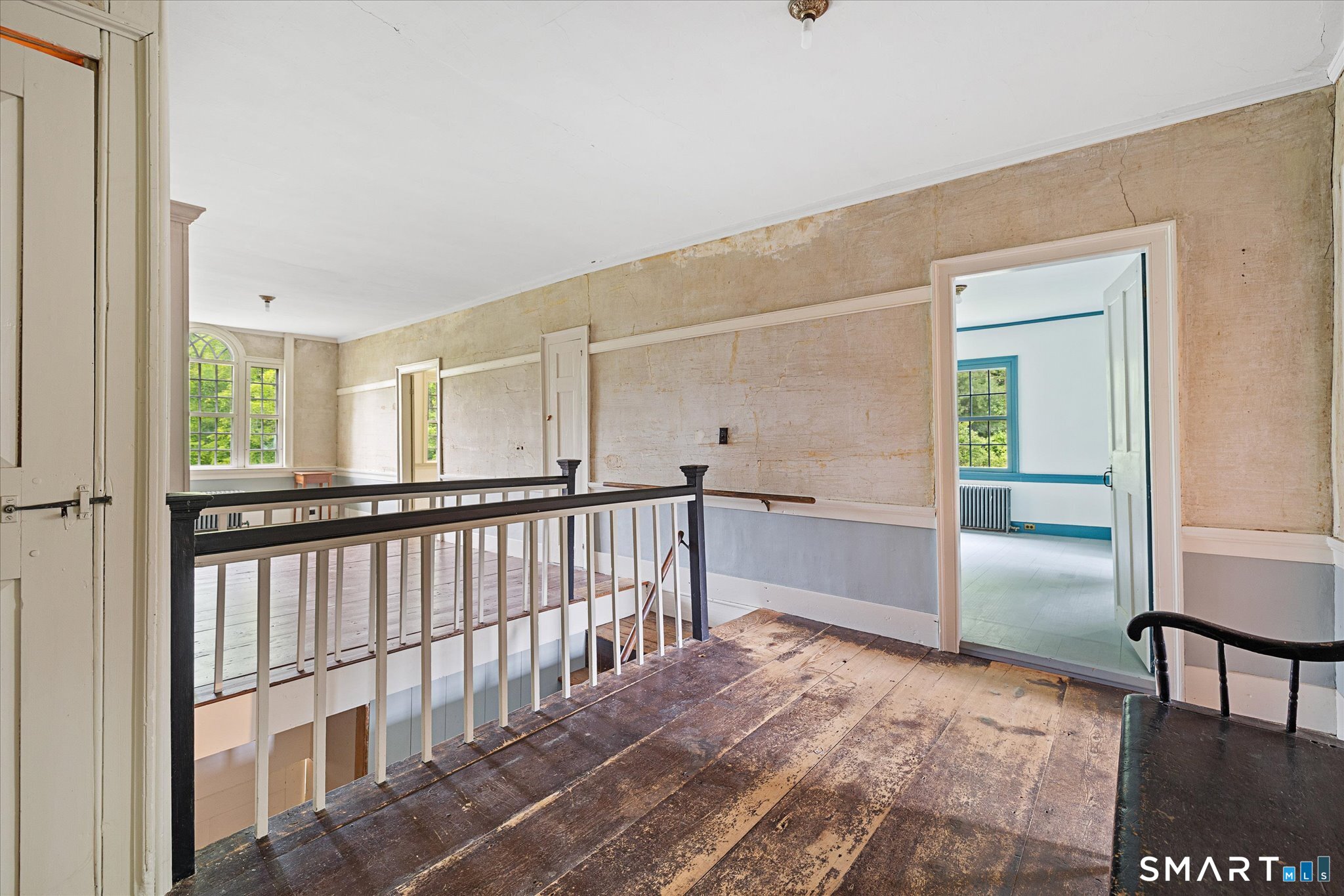Upper-floor landing with worn plank floors, dark stair railing, tall windows, and a blue-trimmed doorway.