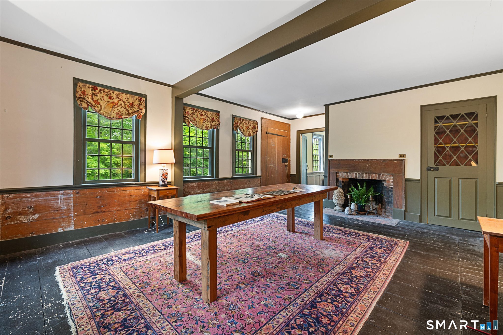Furnished dining room with a long wooden table, brick fireplace, and tall windows.