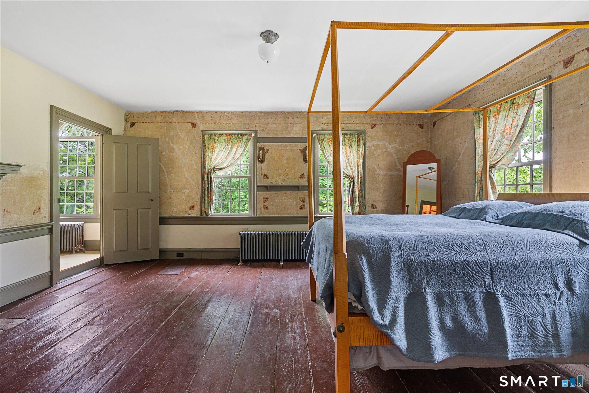 Bedroom with a wooden four-poster bed, blue quilt, worn plank floors, and tall curtained windows.