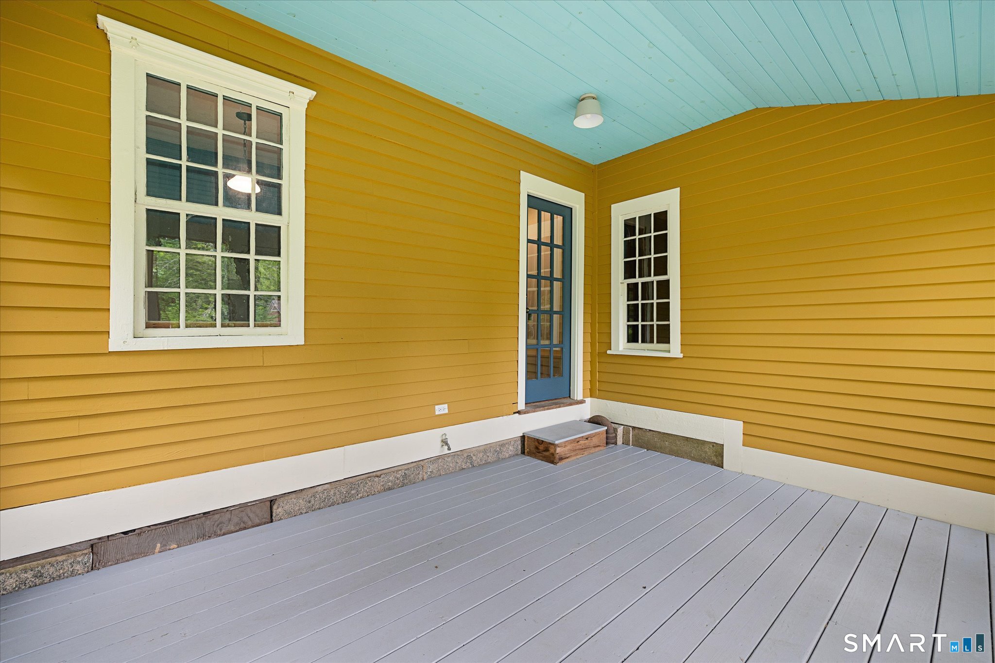 Covered porch with mustard-yellow siding, white-trimmed windows, blue door, and gray decking.