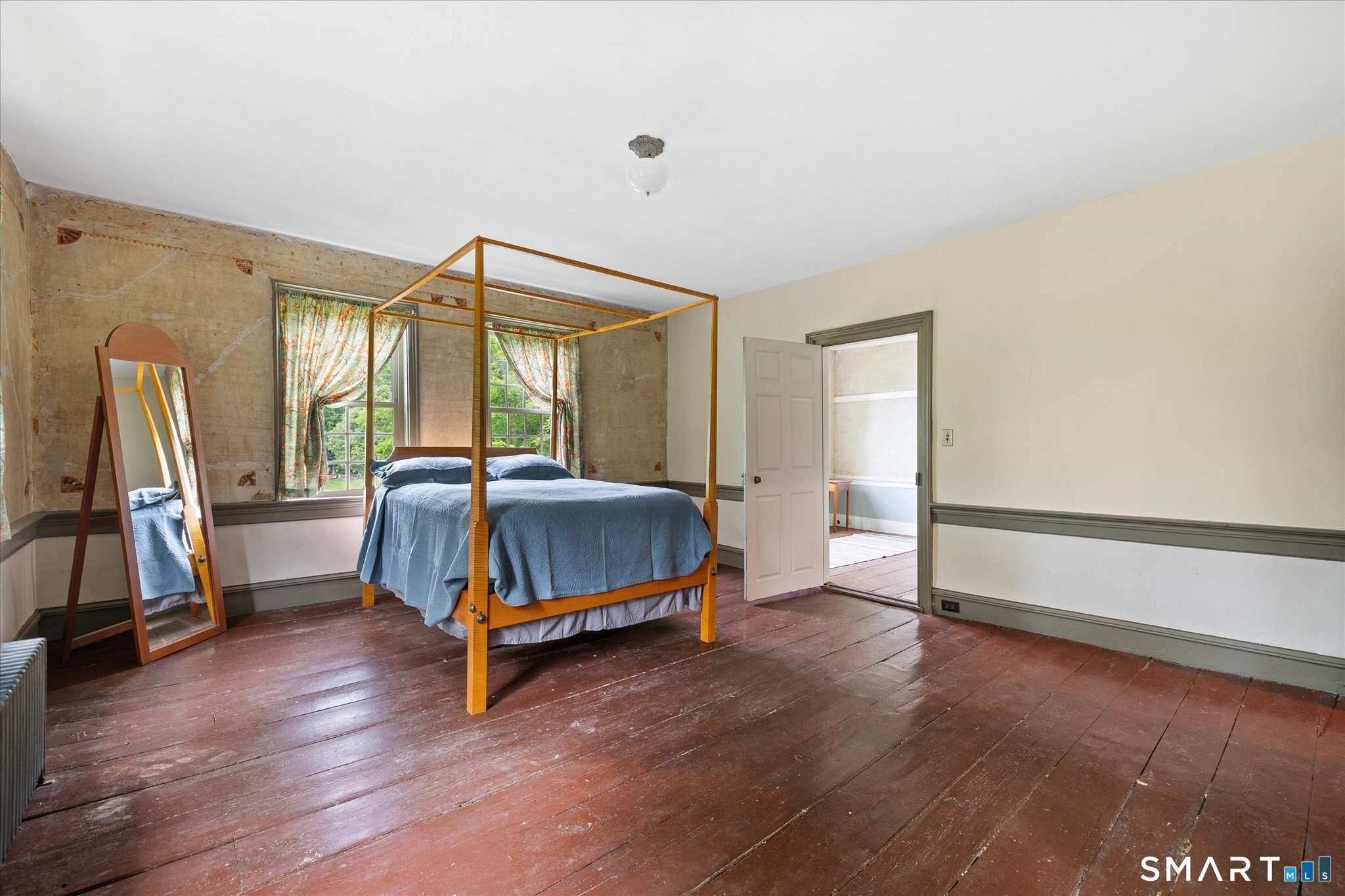 Bedroom with a blue-covered four-poster bed, leaning mirror, and worn wide-plank wood floors.