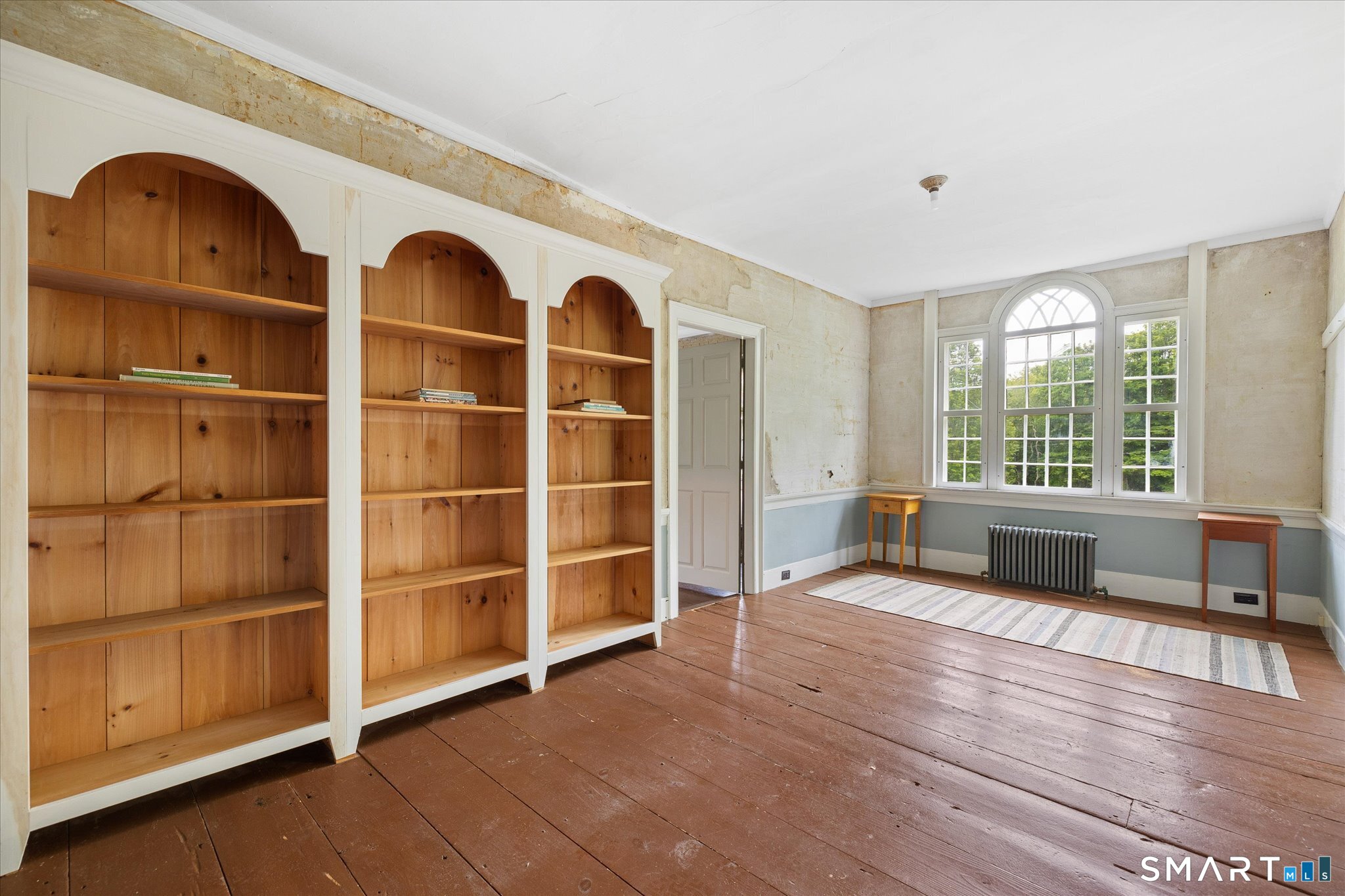 Room with arched window, built-in wooden bookcases, and wide-plank wood floors.