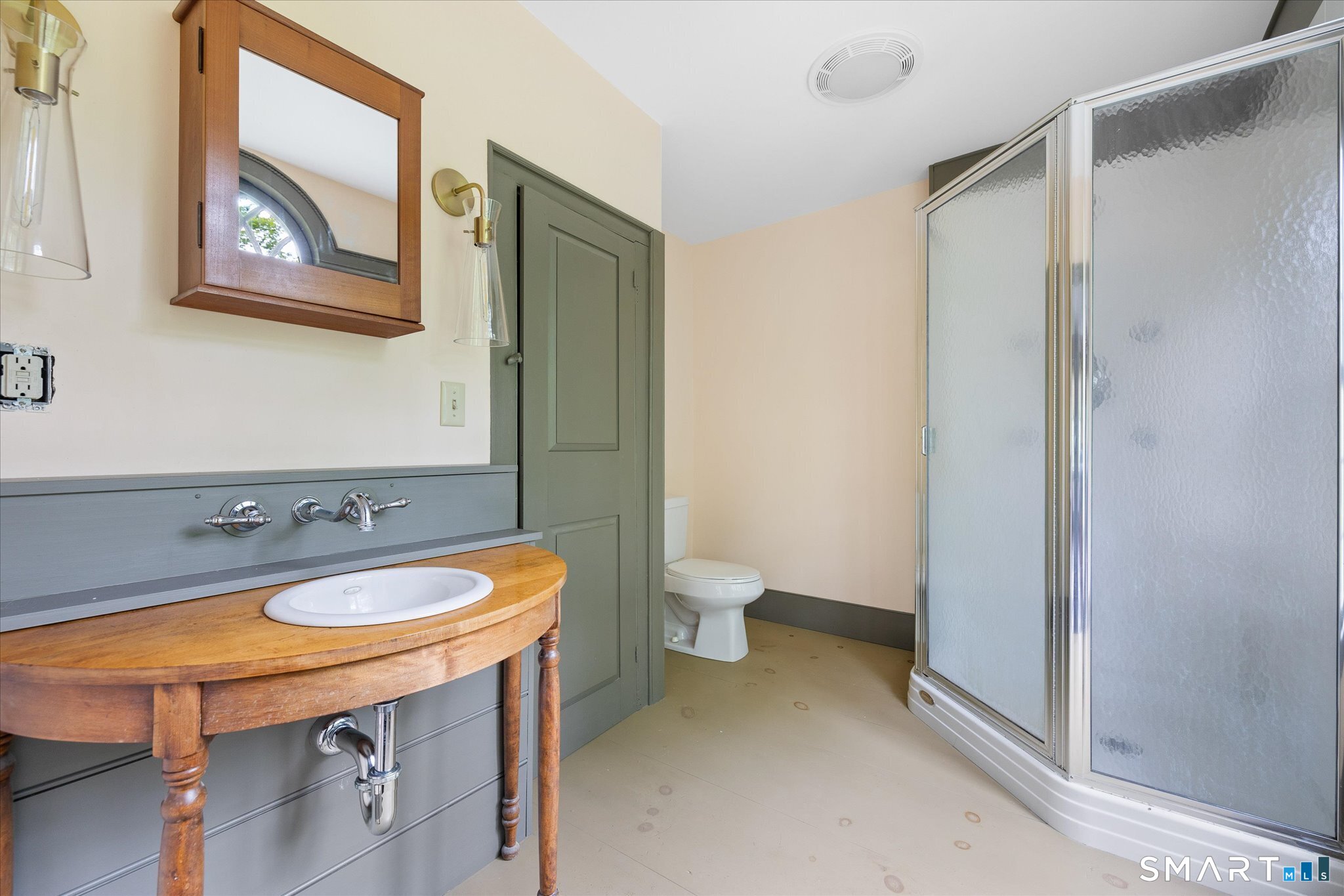 Bathroom with a curved wooden vanity, gray-green door, toilet, and frosted-glass corner shower.