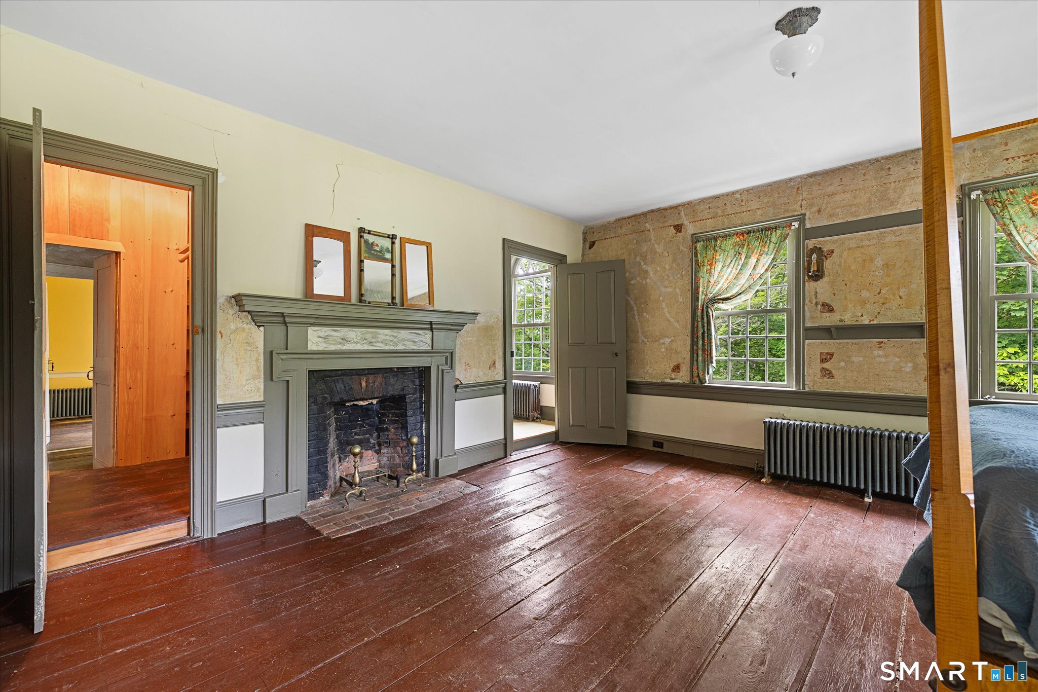 Bedroom with a large fireplace, exposed wall finishes, tall curtained windows, and dark wood floors.