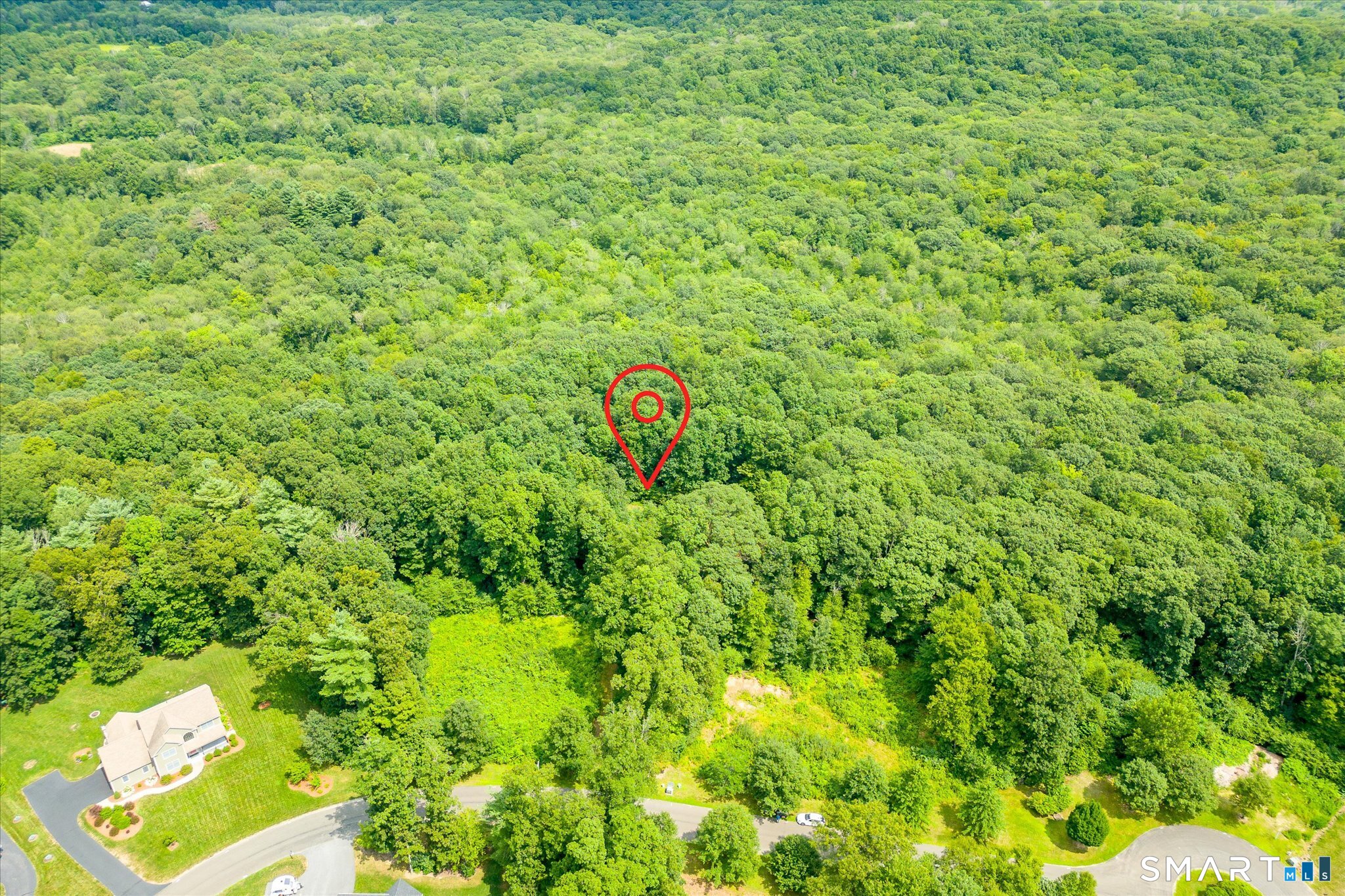 Aerial view of wooded land marked by a red location pin, with a neighboring home and road nearby.