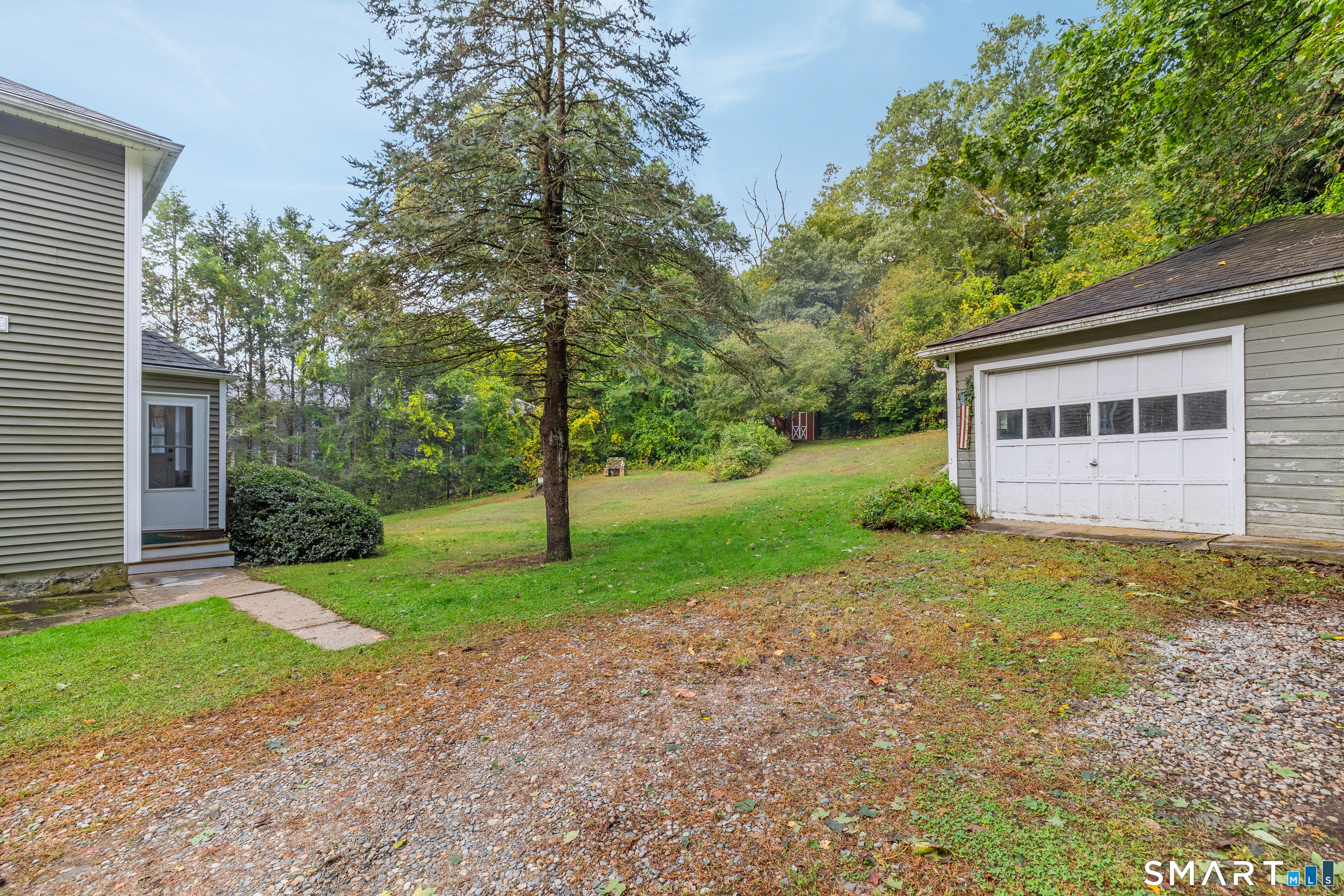 Property yard between the house and detached garage, with a gravel driveway and wooded backdrop.