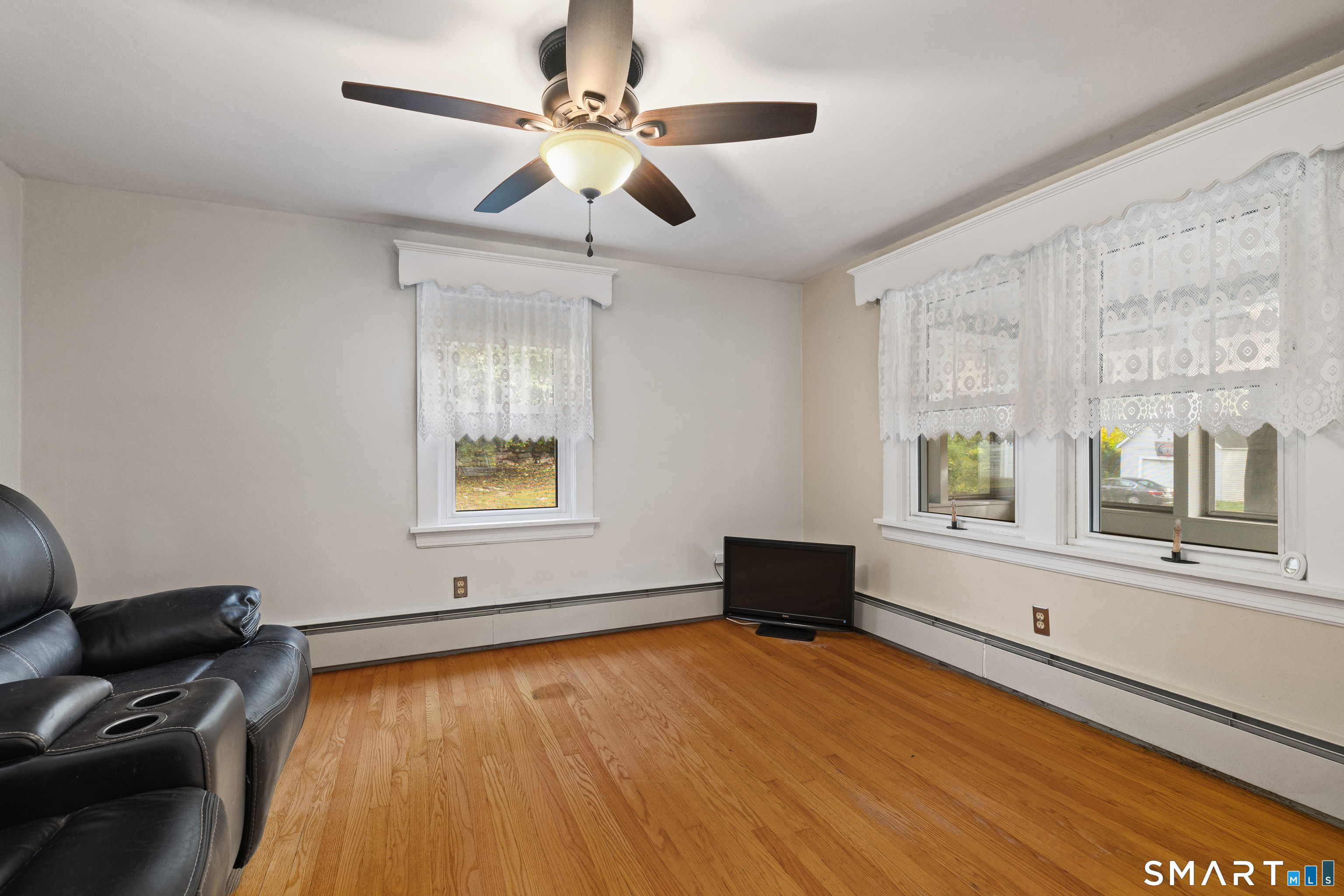 Living room with hardwood floors, lace-curtained windows, a leather recliner, and ceiling fan.