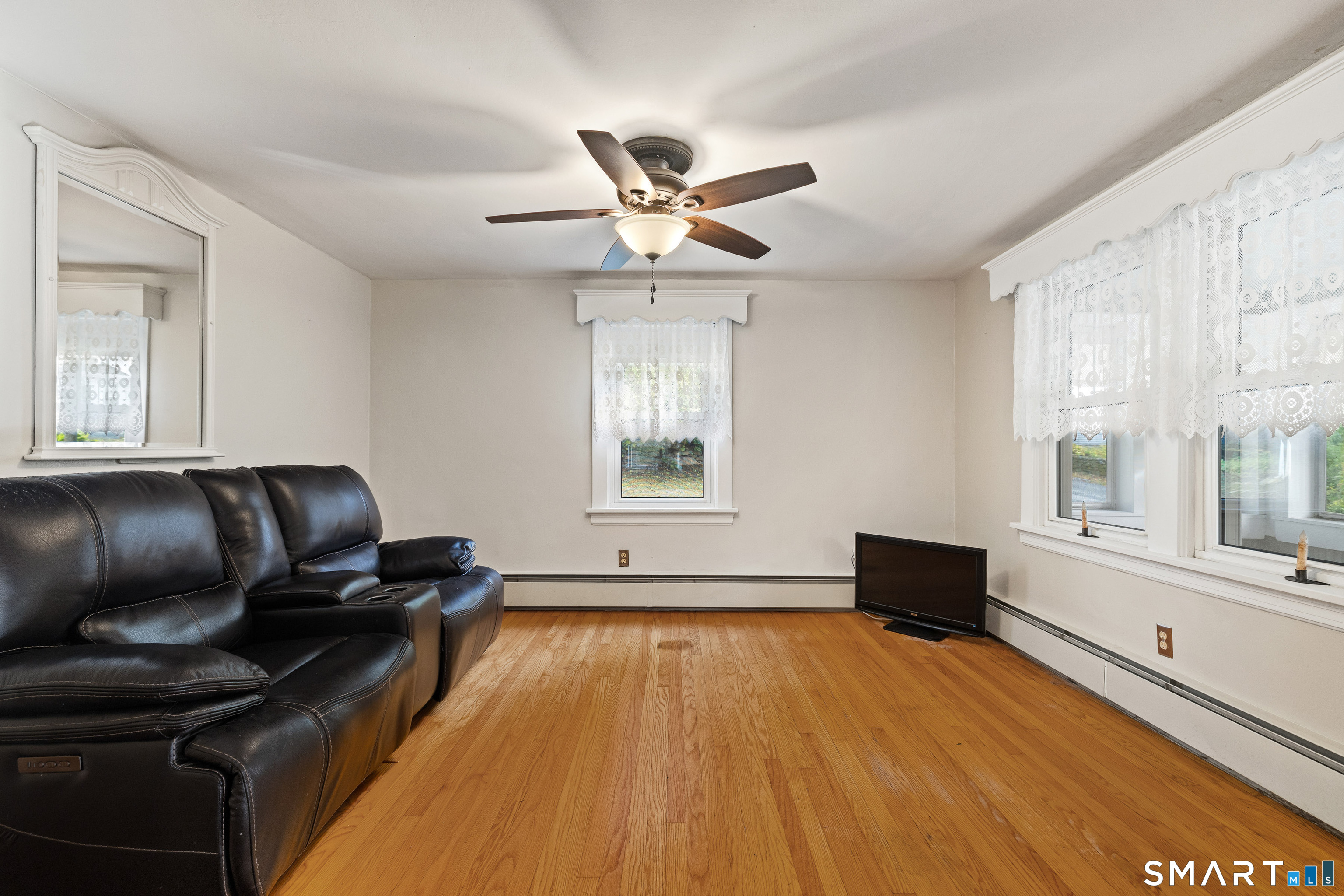 Living room with a black leather reclining sofa, hardwood floors, and lace-curtained windows.