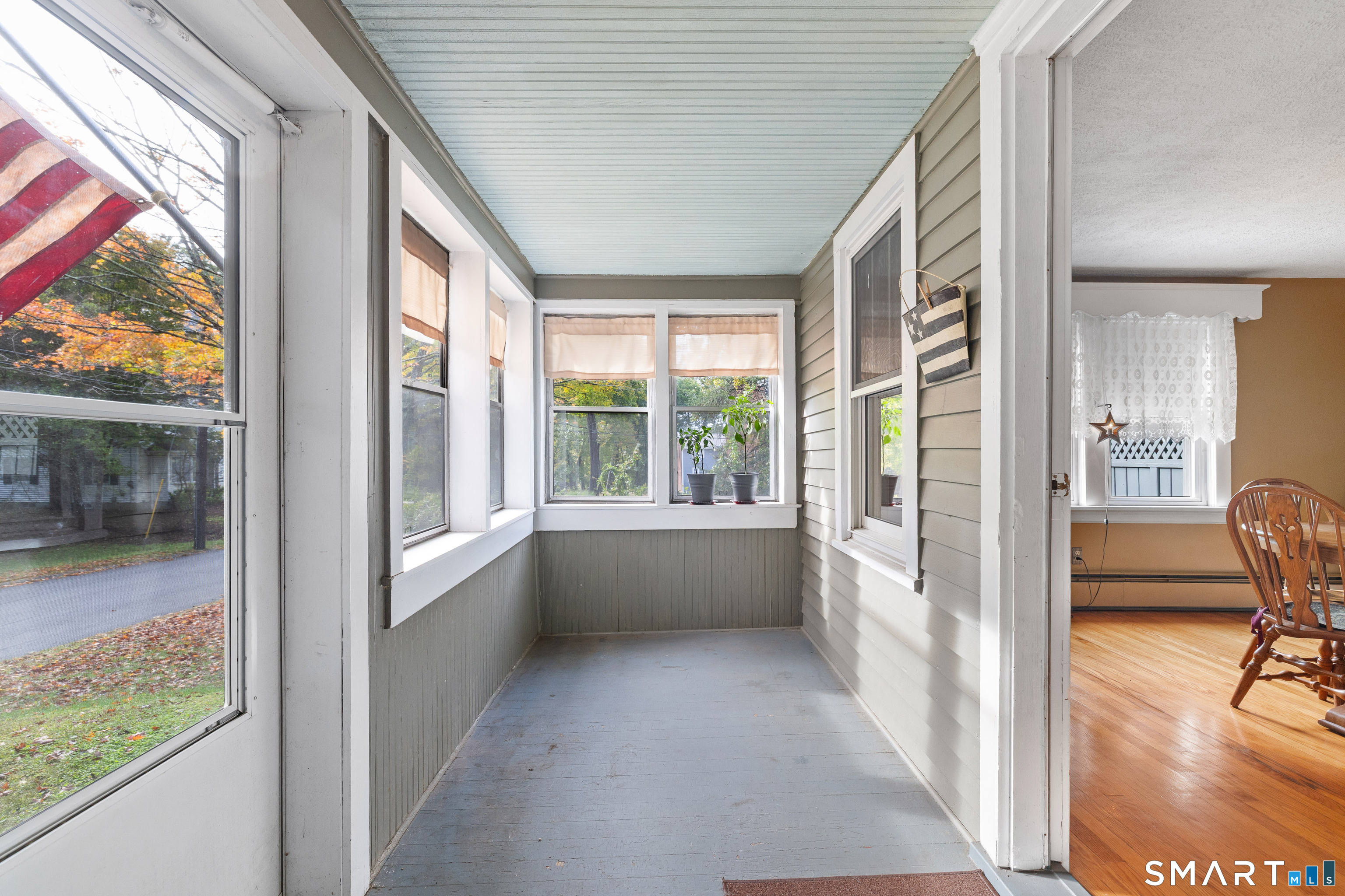 Enclosed porch with many windows, potted plants, and autumn trees visible outside.