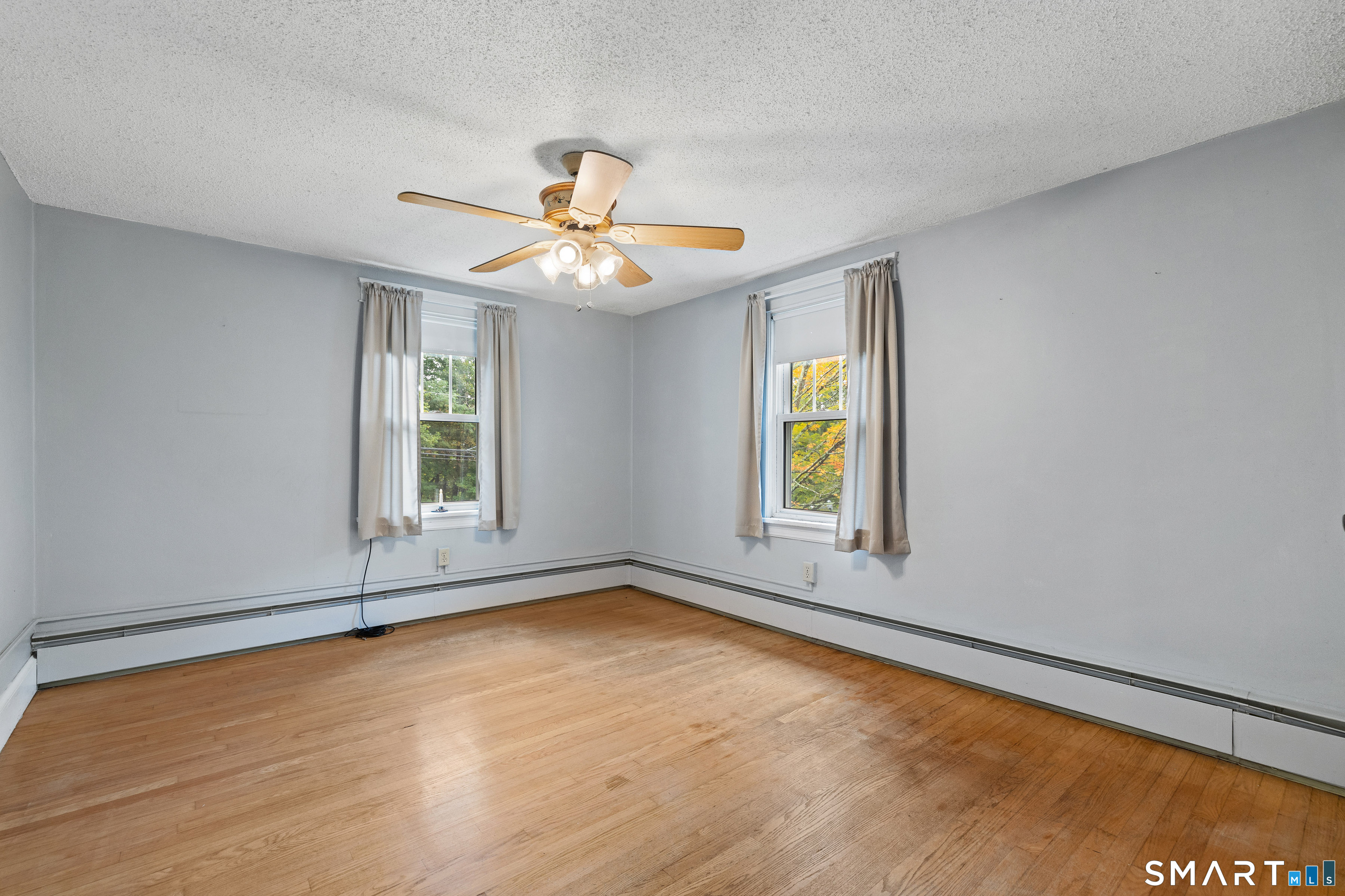 Empty room with hardwood floors, two curtained windows, baseboard heating, and a ceiling fan.
