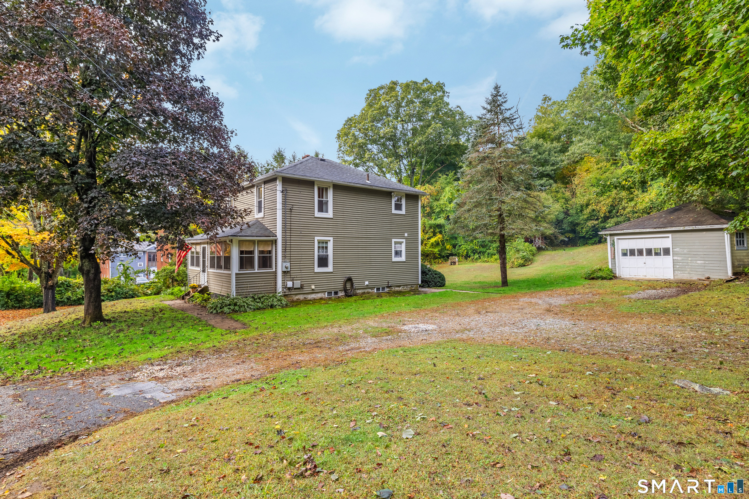 Two-story taupe house and detached garage amid autumn trees, with a gravel driveway and broad lawn.