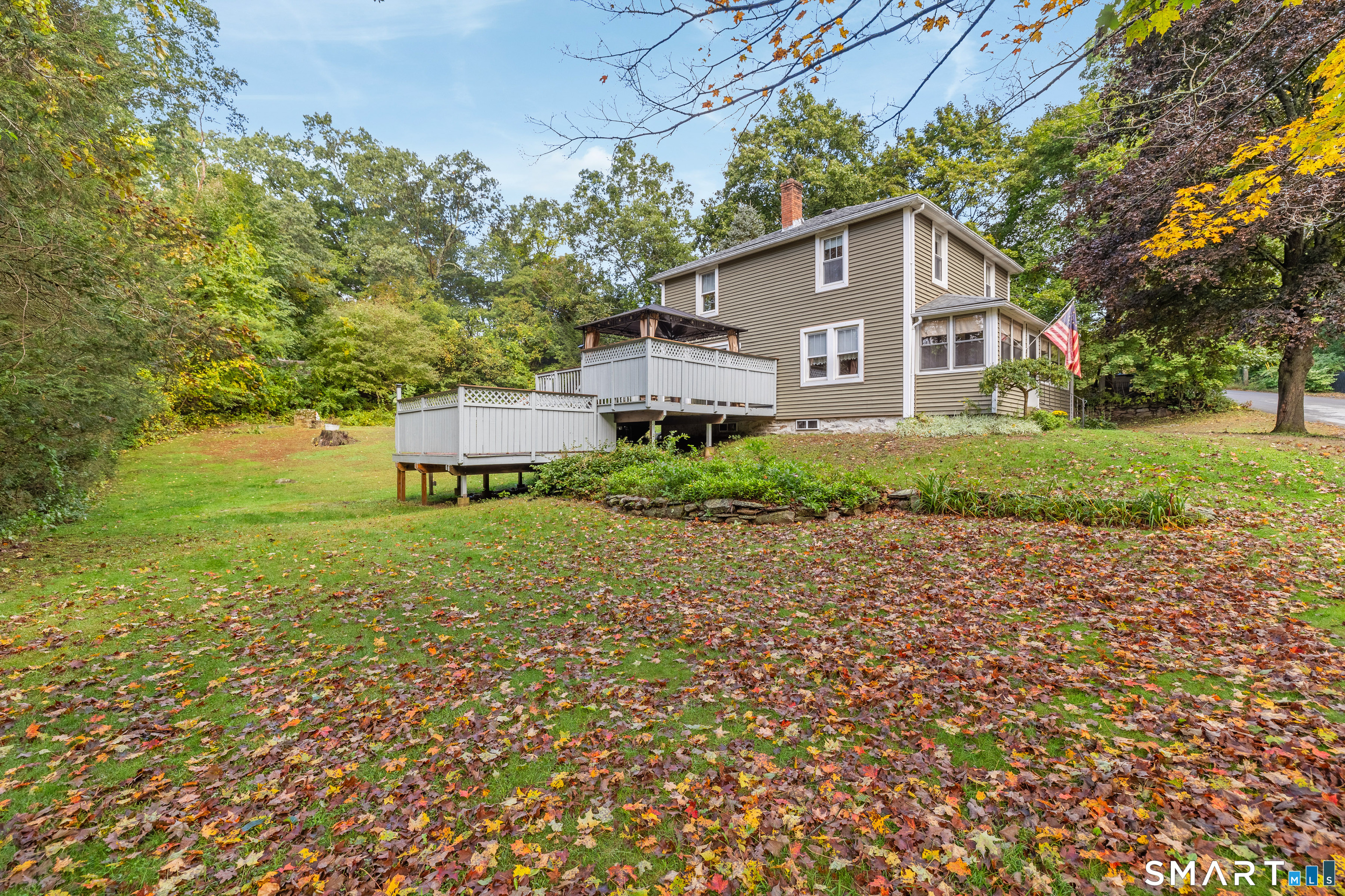 Two-story tan house with elevated white-railed decks and a leaf-covered lawn in autumn.