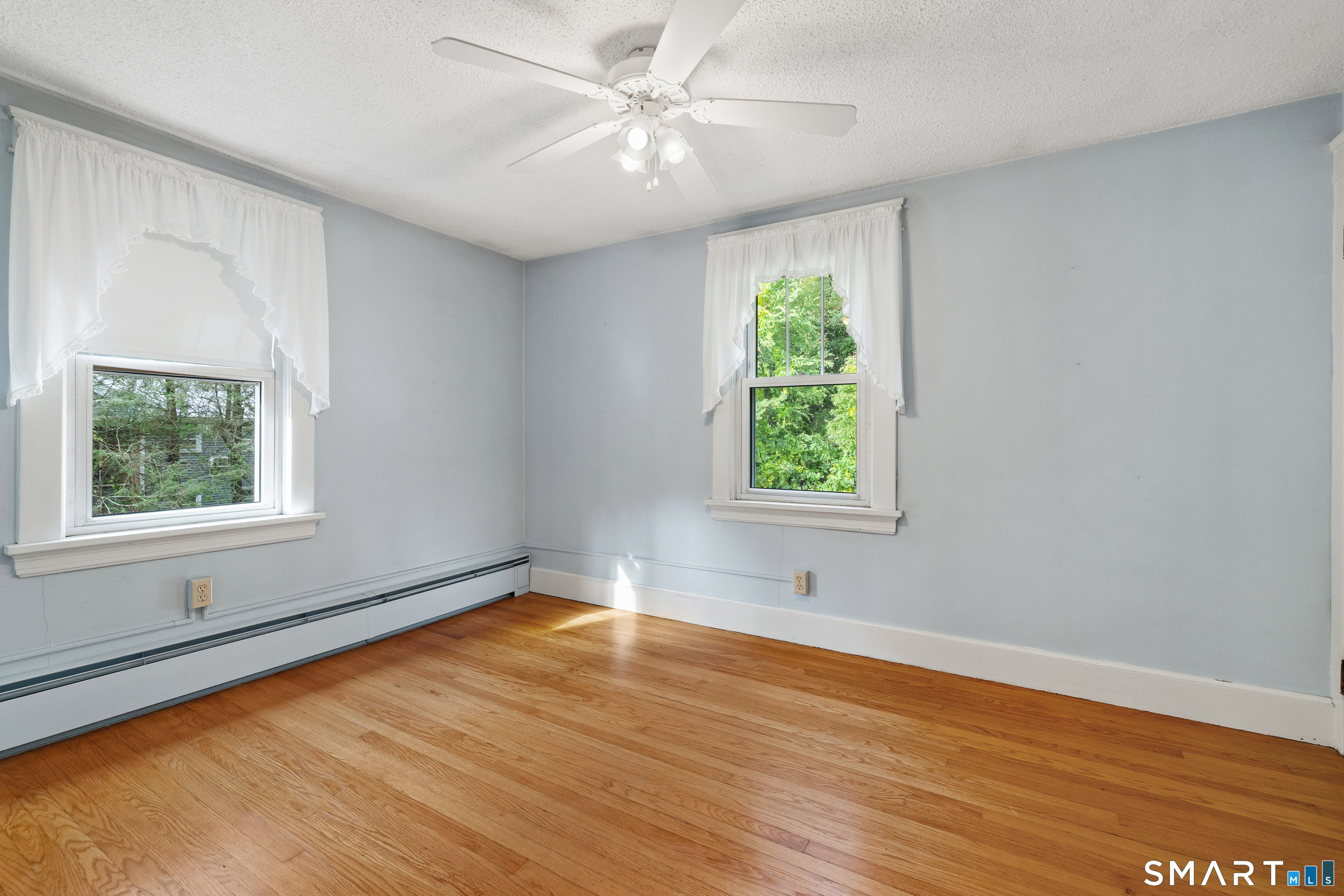 Vacant room with hardwood floors, two curtained windows, and a ceiling fan.