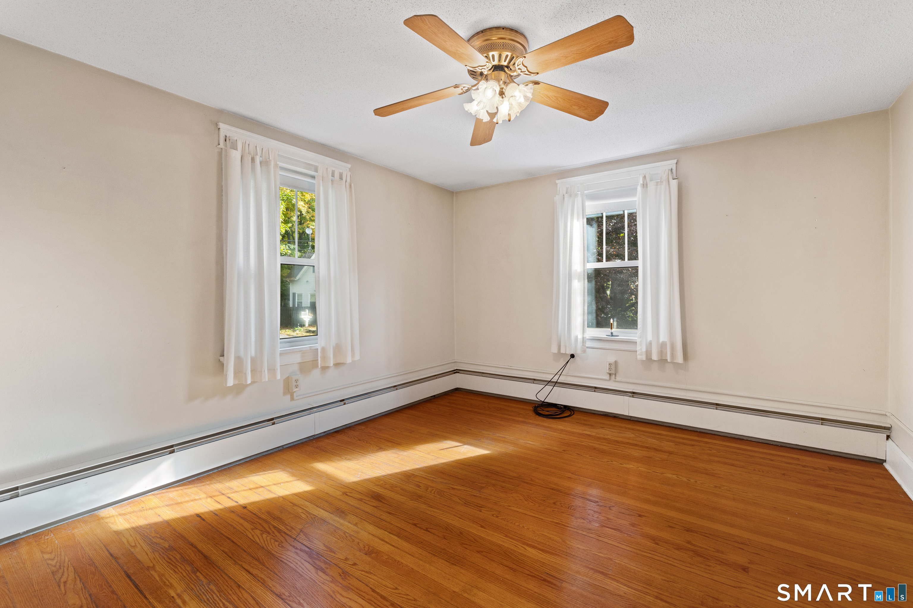 Empty room with polished hardwood floors, two curtained windows, and a ceiling fan.