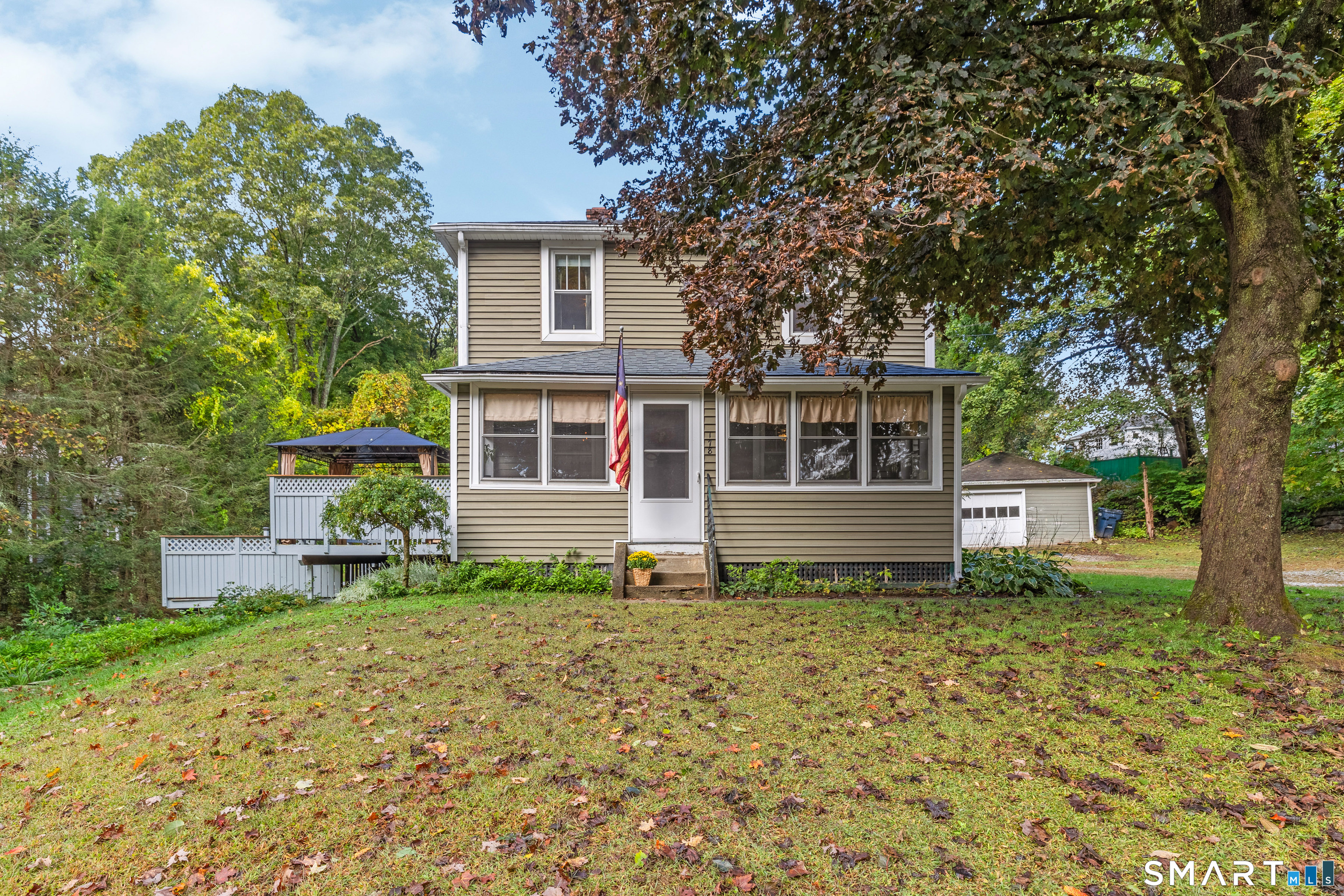 Two-story beige house with a screened porch, white deck, and mature trees in an autumn yard.