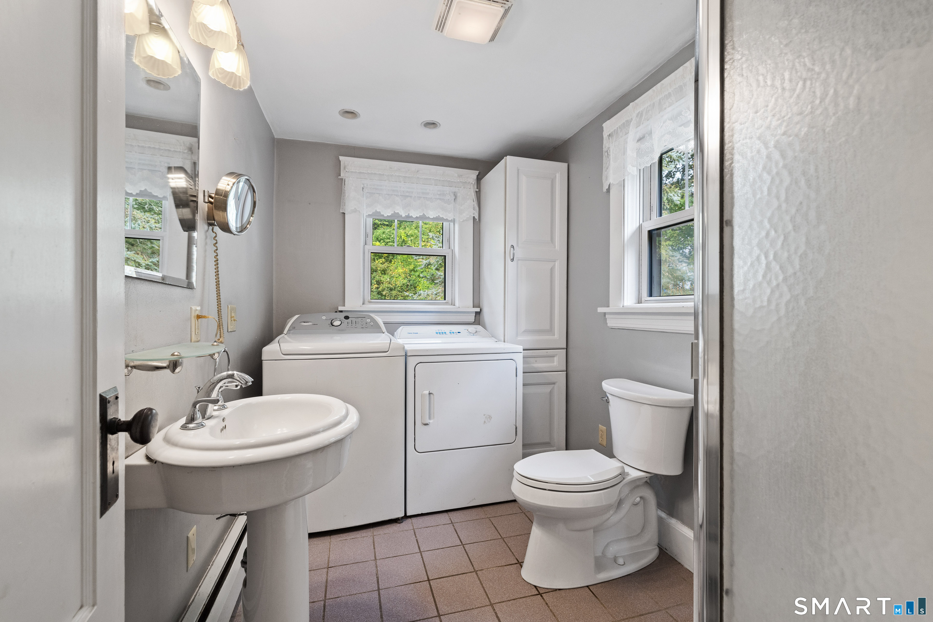 Bathroom and laundry room with a pedestal sink, toilet, washer, dryer, and two windows.