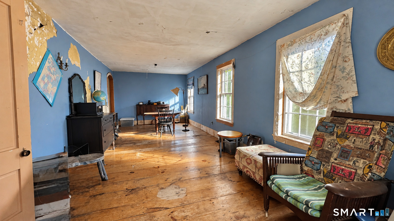 Furnished room with blue walls, worn wood floors, tall windows, and peeling paint on the walls and ceiling.
