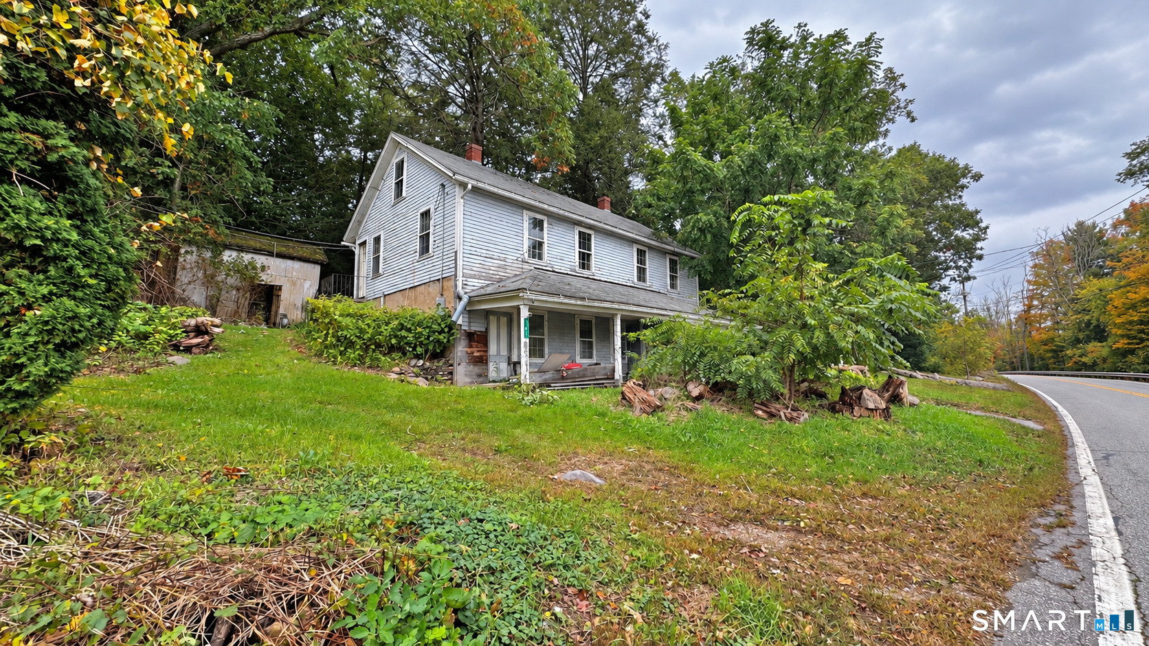 Two-story white clapboard house on an overgrown slope beside a curving road.